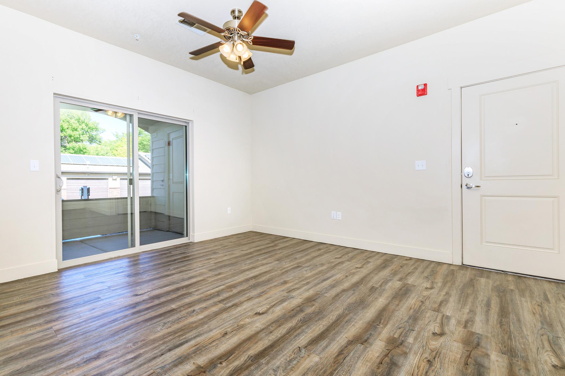 A spacious, empty room with light-colored walls and a ceiling fan. There is a sliding glass door leading to a balcony, and a front door on the right. The floor is a warm, wooden laminate, creating a bright and open atmosphere. Natural light illuminates the space from the door.