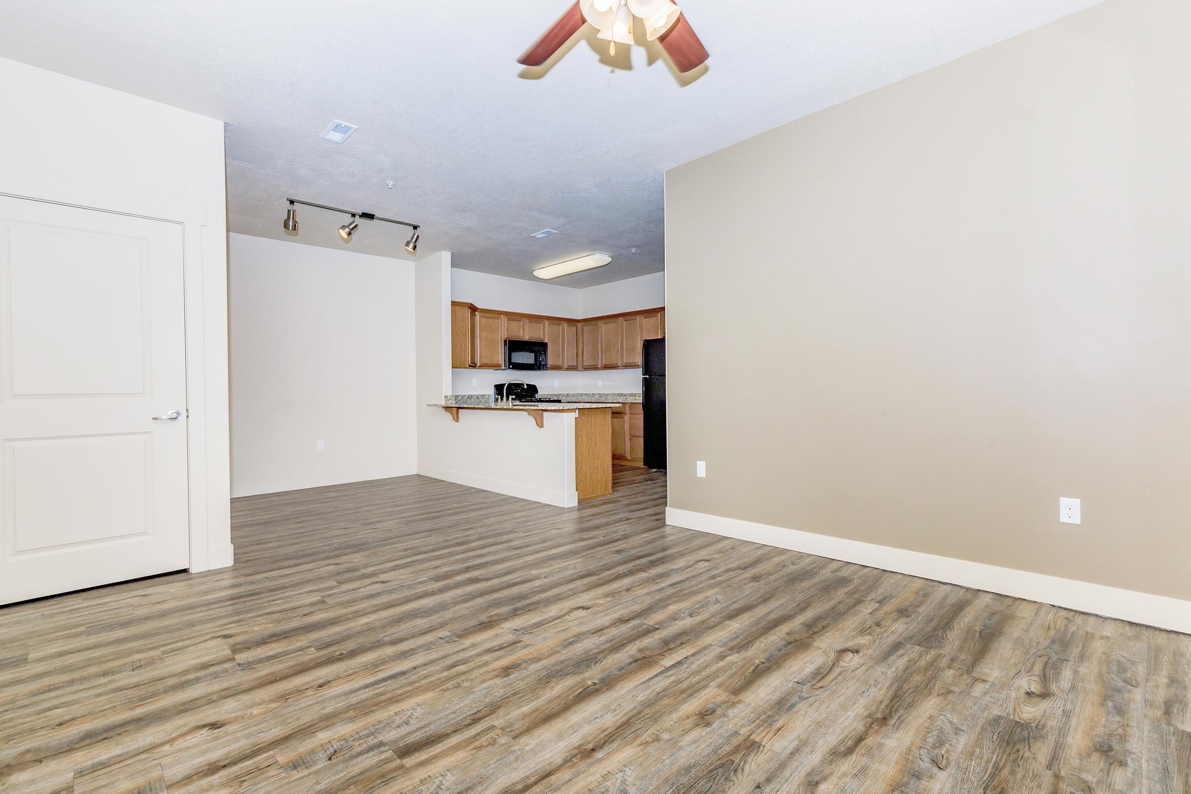 A spacious, modern living room with light brown walls and wooden flooring. It features a ceiling fan and is open to a kitchen area in the background, which includes wood cabinets and kitchen appliances. Natural light fills the space, creating a welcoming atmosphere.