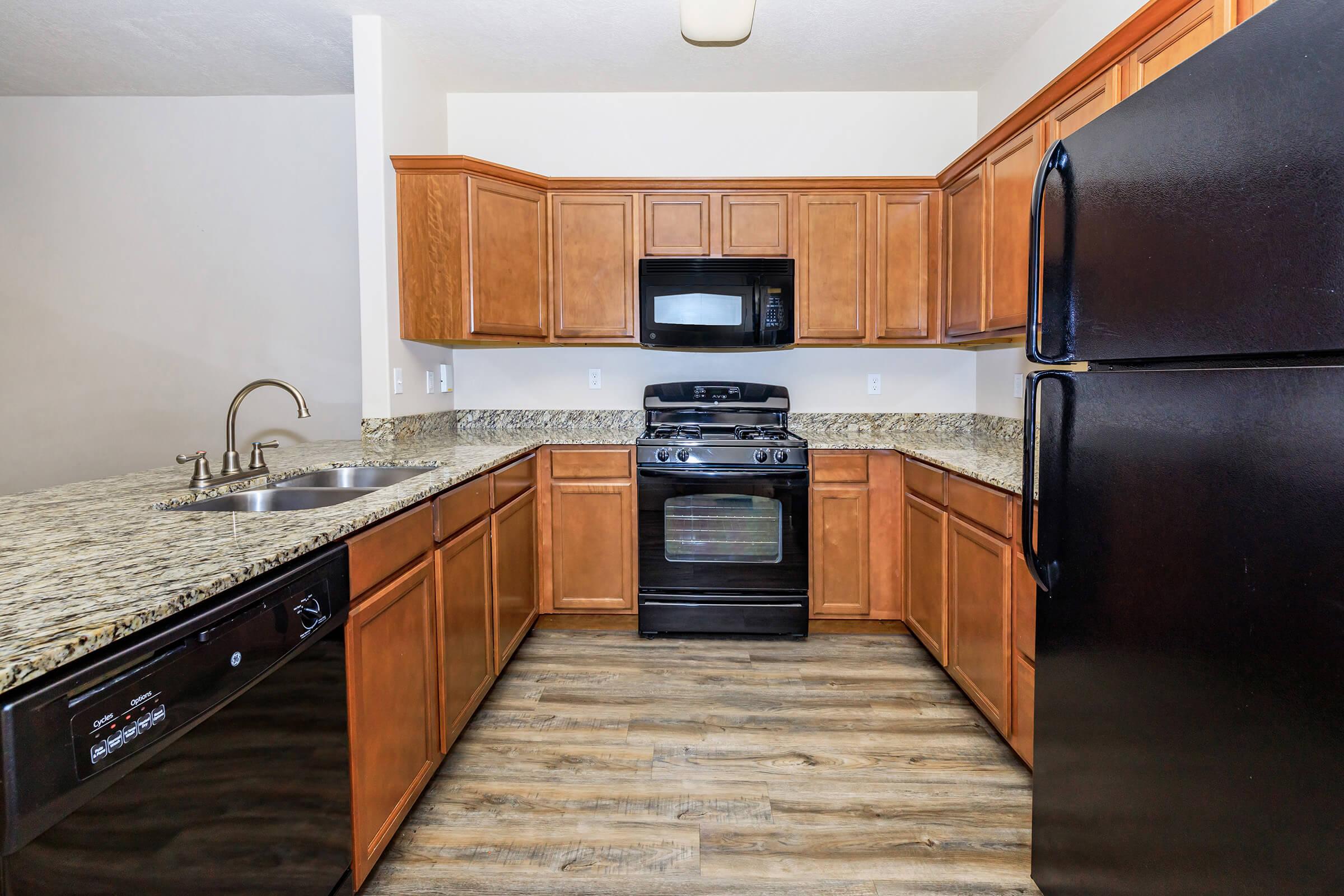 A modern kitchen featuring wooden cabinets, a granite countertop, stainless steel sink, and black appliances, including a stove, microwave, and refrigerator. The space has a clean and organized layout with wooden flooring and ample natural light. 