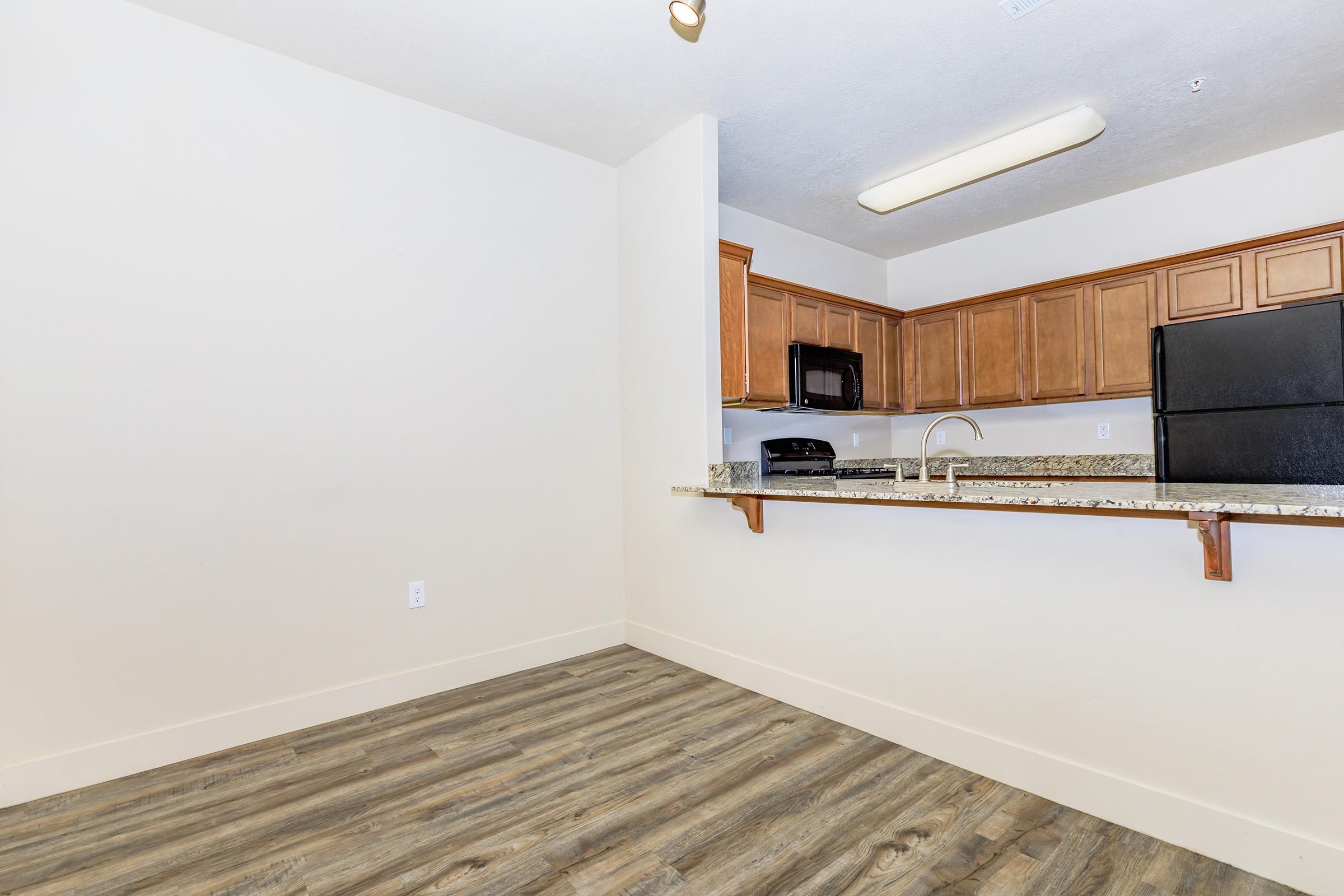 Interior view of a modern kitchen with wooden cabinetry and granite countertops. Includes a black microwave and refrigerator. The flooring is light brown, and the walls are painted a neutral color. The layout features an open space connecting the kitchen to the living area.