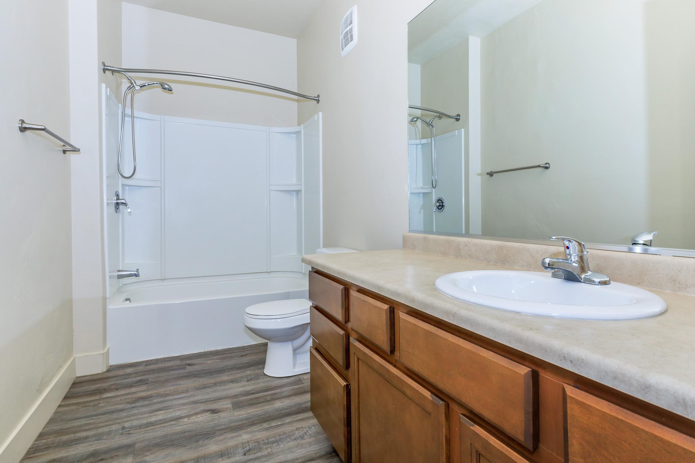 A clean and modern bathroom featuring a bathtub with a shower, a toilet, and a wooden vanity with a sink. The walls are neutral-colored, and there are mirrors on the wall reflecting the space. The flooring is a wood-like material, giving the room a warm and inviting feel.