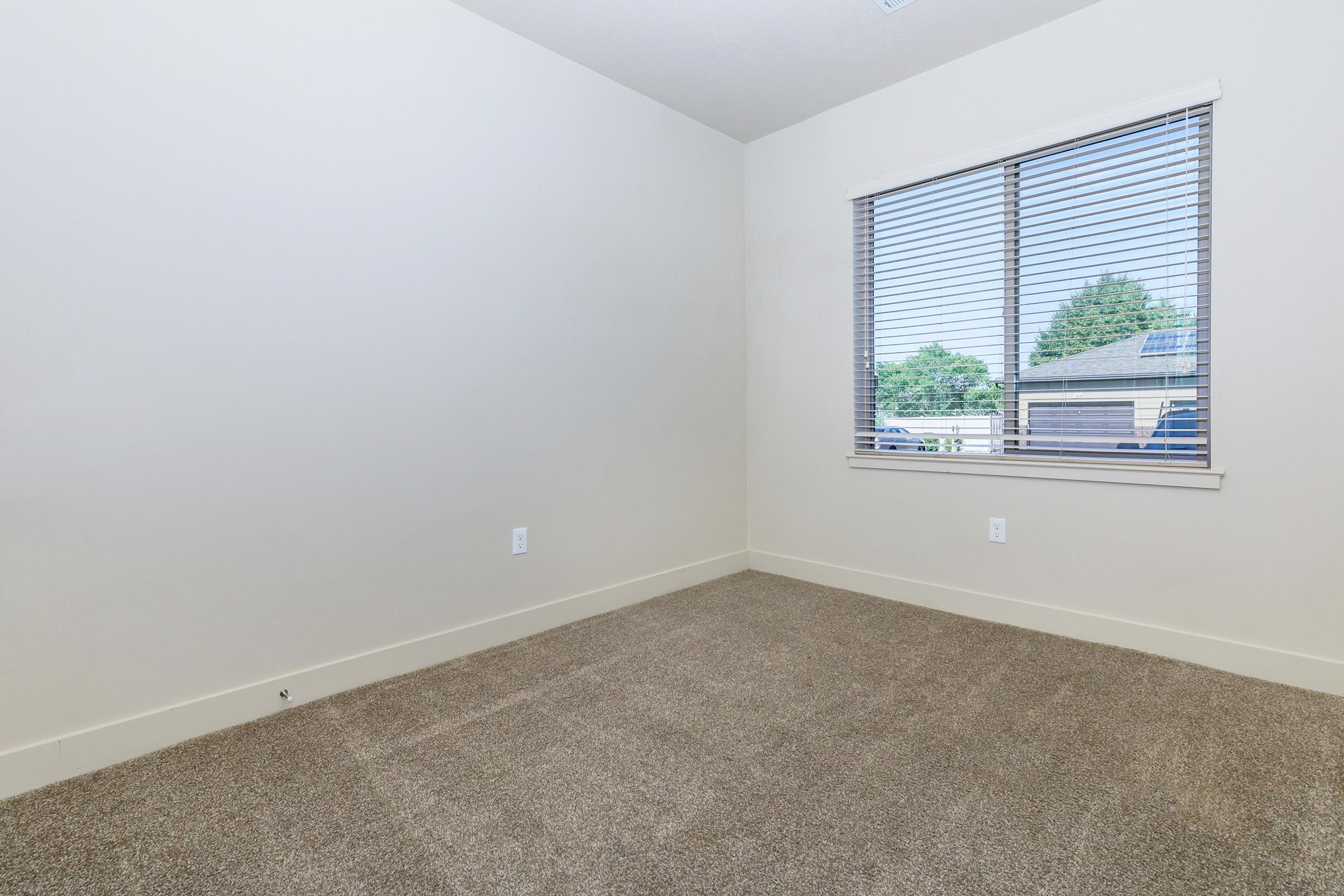 A sparsely furnished room with light beige walls and a large window featuring white blinds. The floor is covered in brown carpet. Sunlight streams in through the window, illuminating the empty space. The room has a minimalistic appearance, making it suitable for various purposes.