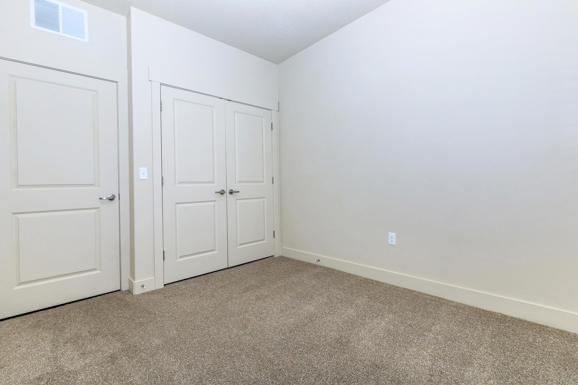 A brightly lit room featuring light-colored walls and carpet. There are two closed double doors on the right side, likely leading to a closet or another space. The left side shows a single door, and the overall atmosphere is clean and minimalistic, emphasizing the spaciousness of the area.