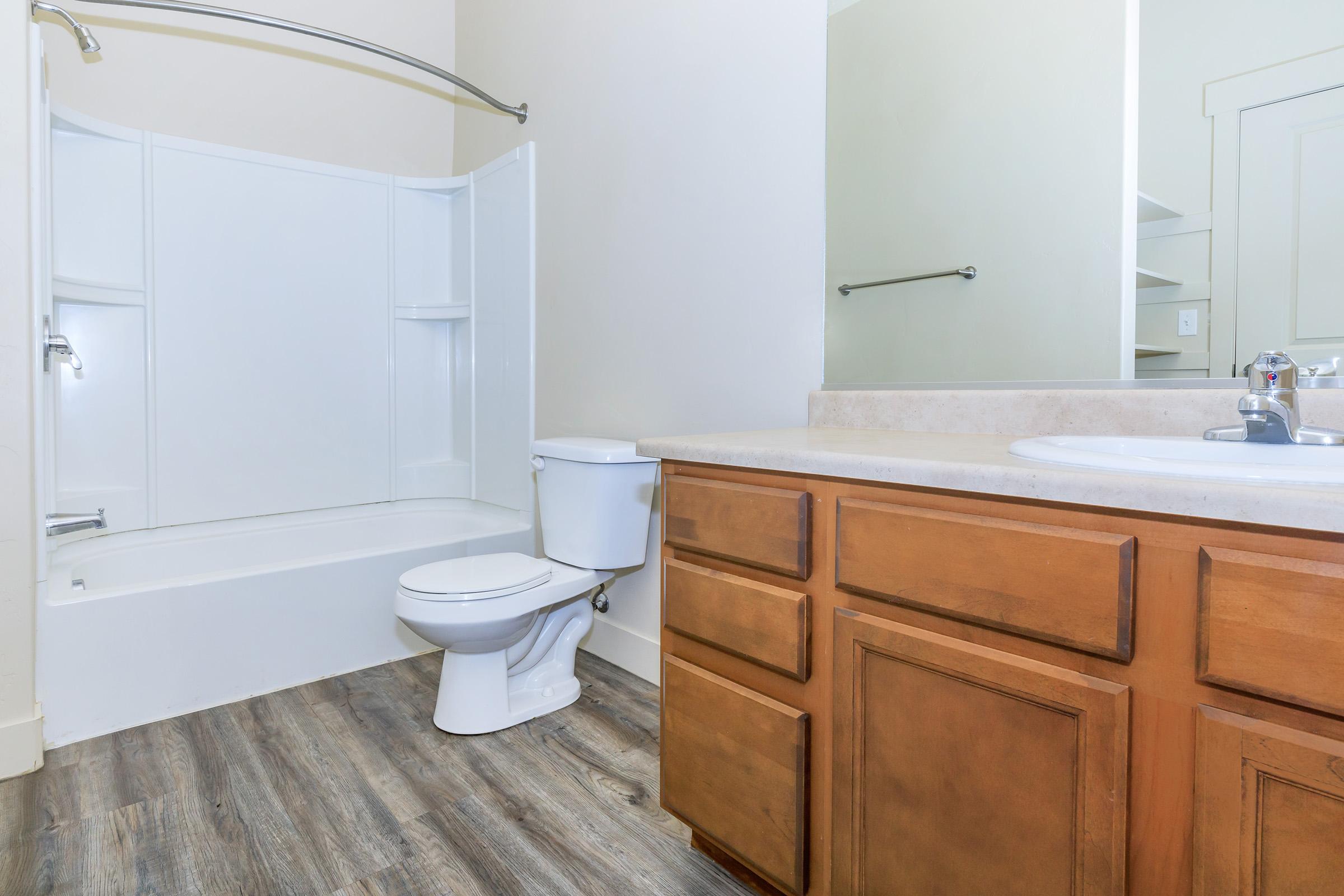 A modern bathroom featuring a white tub and shower unit, a white toilet, and a wooden vanity with a sink. The floor is a light-colored laminate. A mirror is visible above the sink, along with shelving in the corner. The walls are painted in soft, neutral tones.