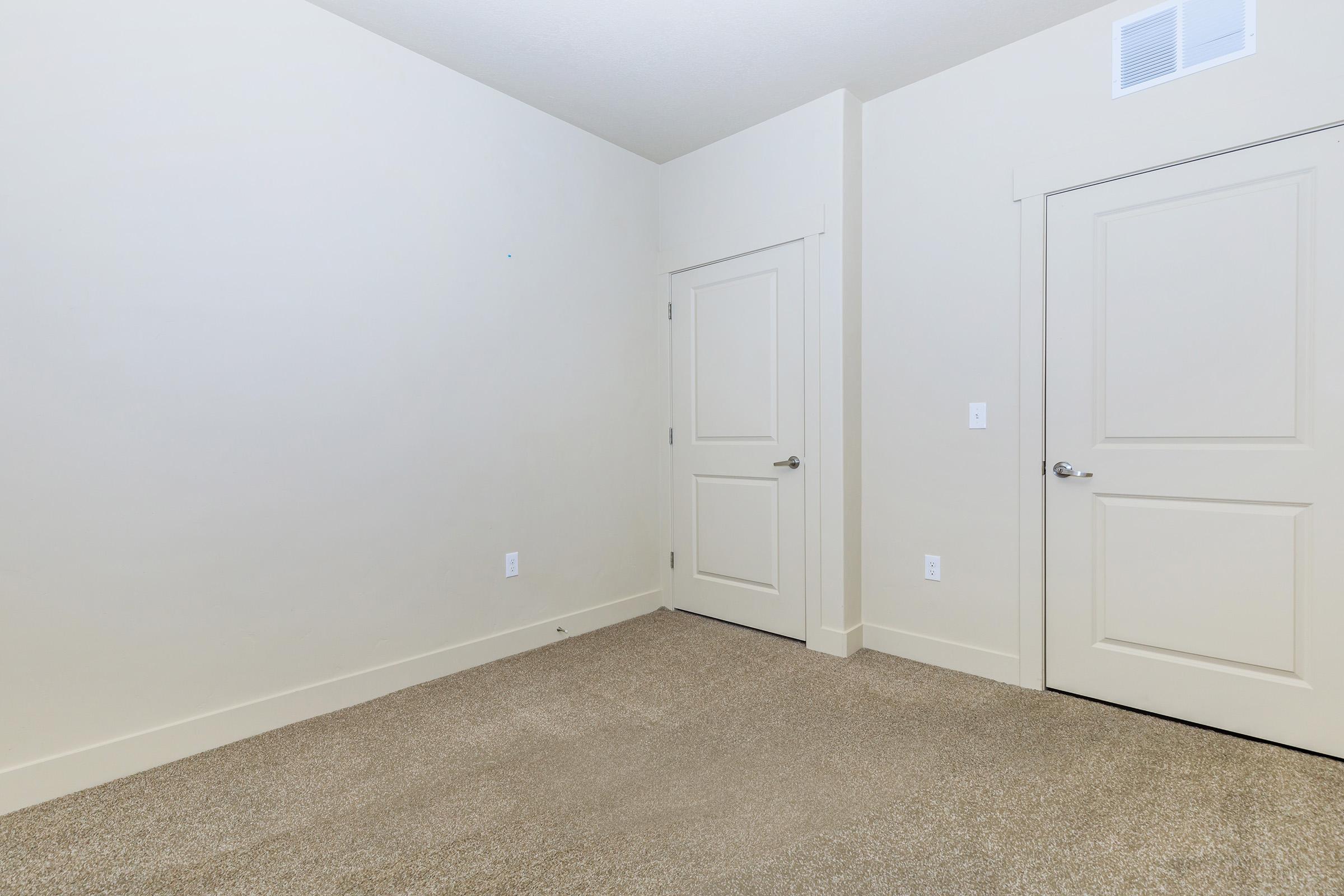 Empty room with light-colored walls, carpeted floor, and a single door on the right. A second door is visible in the background, creating a minimalist and spacious feel. The room has no furniture or decorations, emphasizing its vacant state. Natural light may be present from a nearby window.