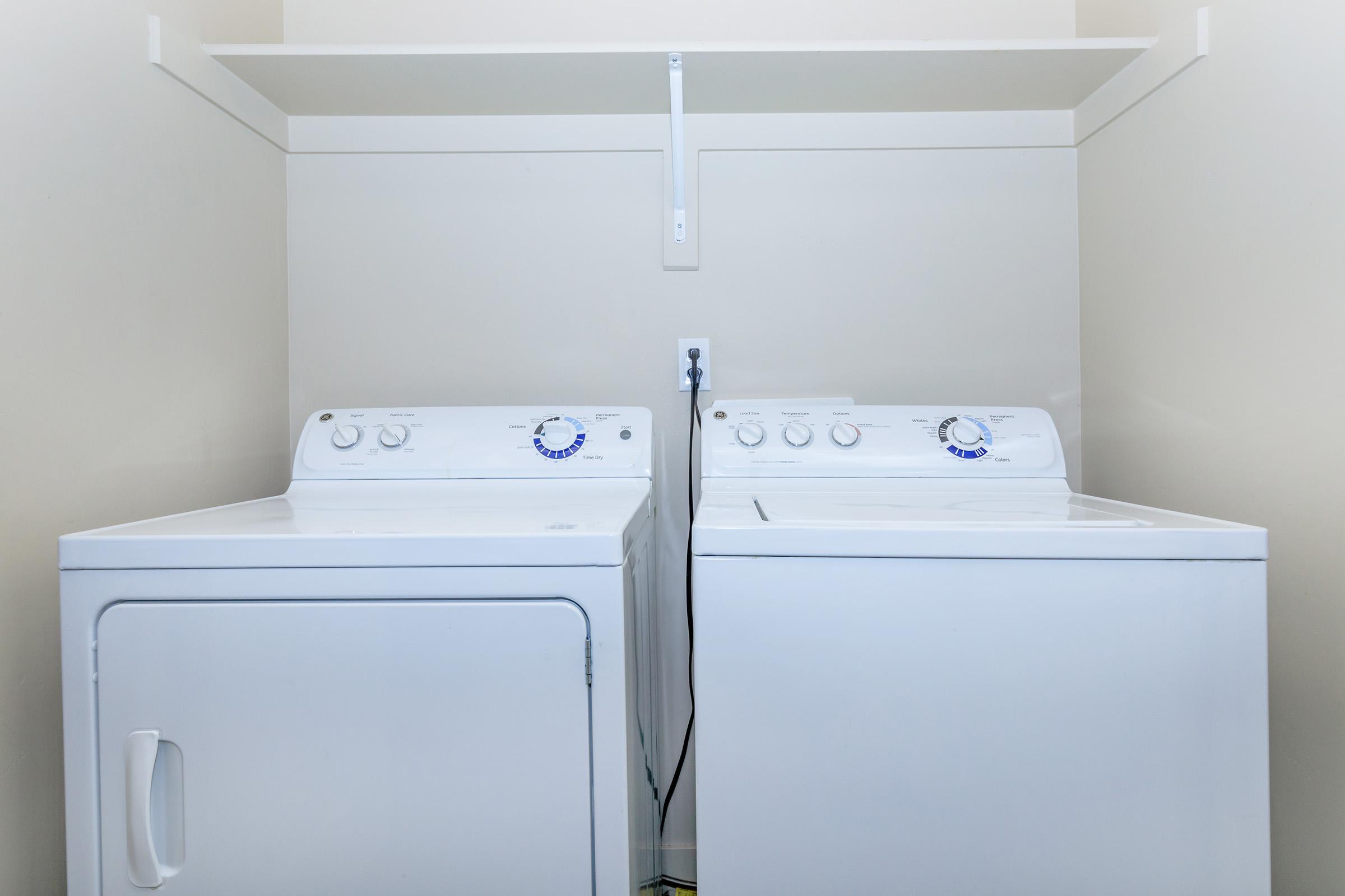 A white washing machine and dryer are side by side in a light-colored laundry space. Above them is a simple shelf mounted on the wall, providing additional storage or display space. The appliances have control dials on the front for easy operation.