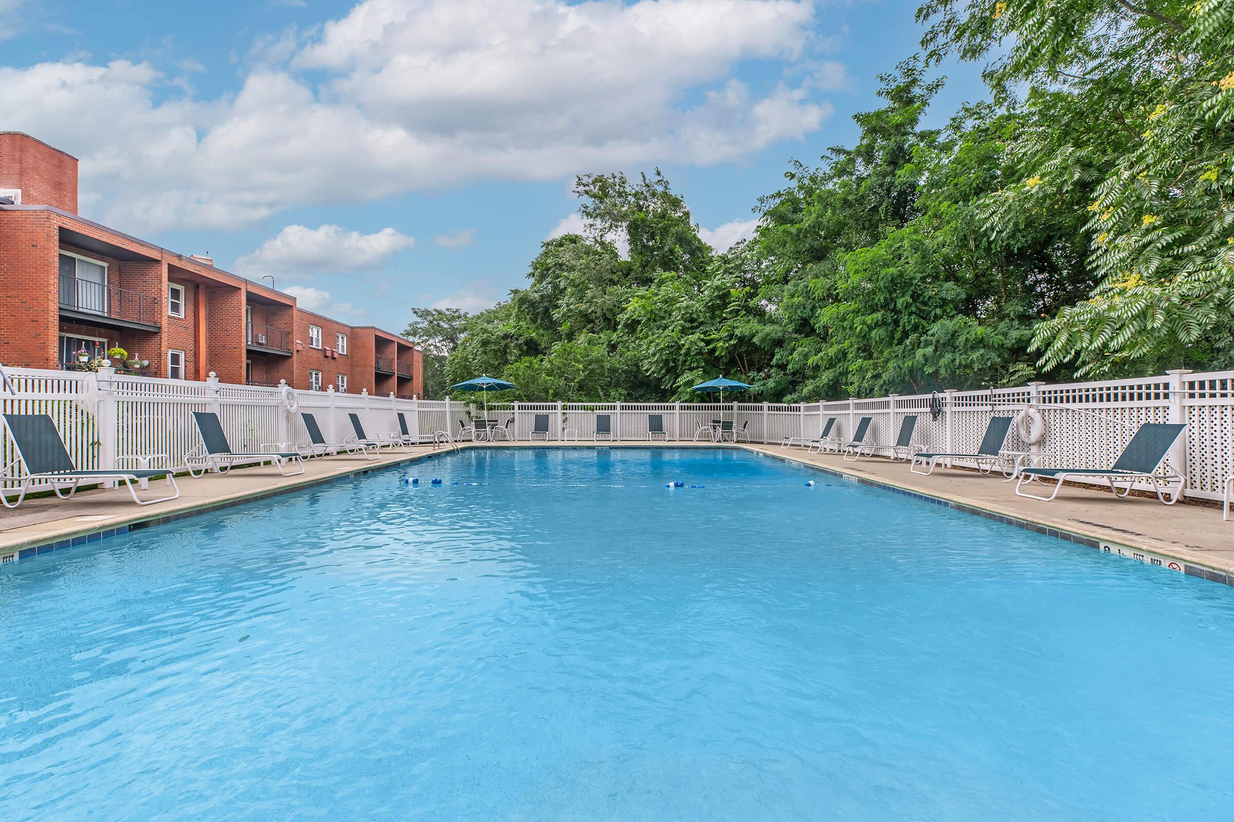 A clear swimming pool surrounded by a white fence, with several lounge chairs arranged around it. Lush green trees and shrubs line the background, under a bright blue sky with fluffy white clouds.