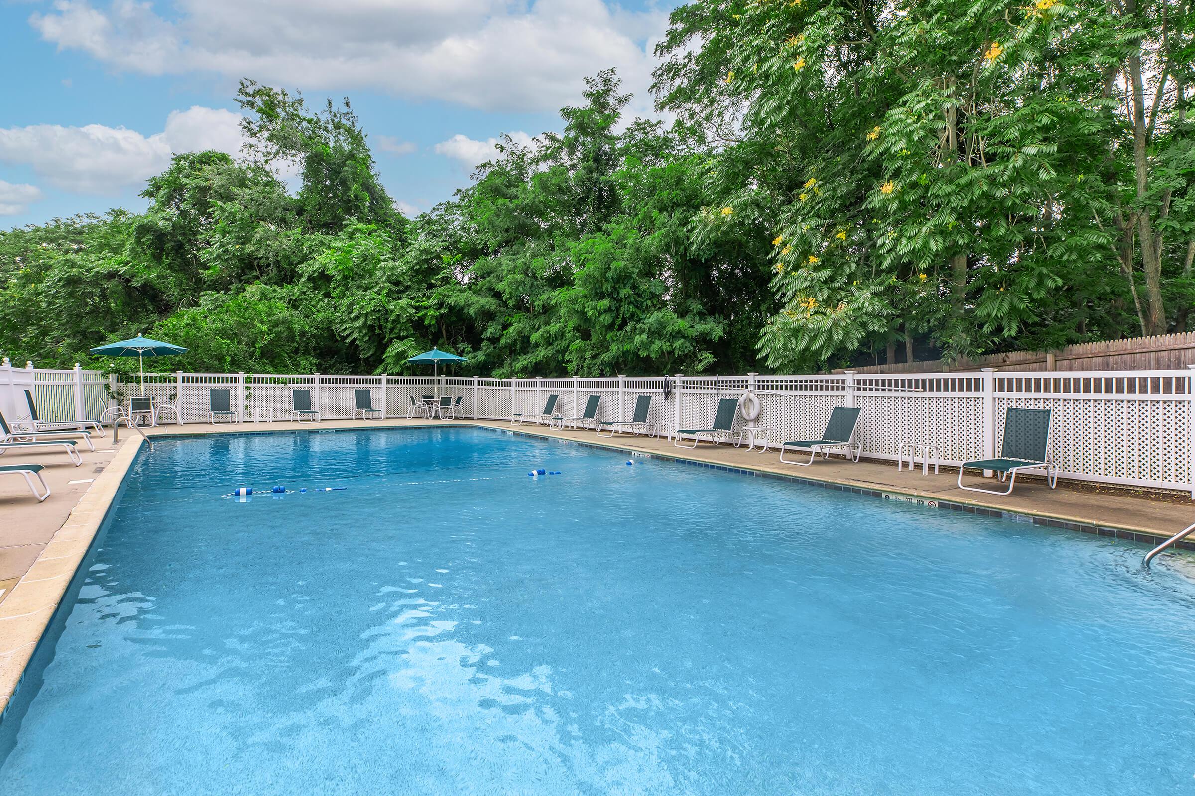 A clear, inviting swimming pool surrounded by a spacious deck. Wooden lounge chairs are arranged along the edge, and green umbrellas provide shade. Lush trees in the background enhance the serene atmosphere under a partly cloudy sky.