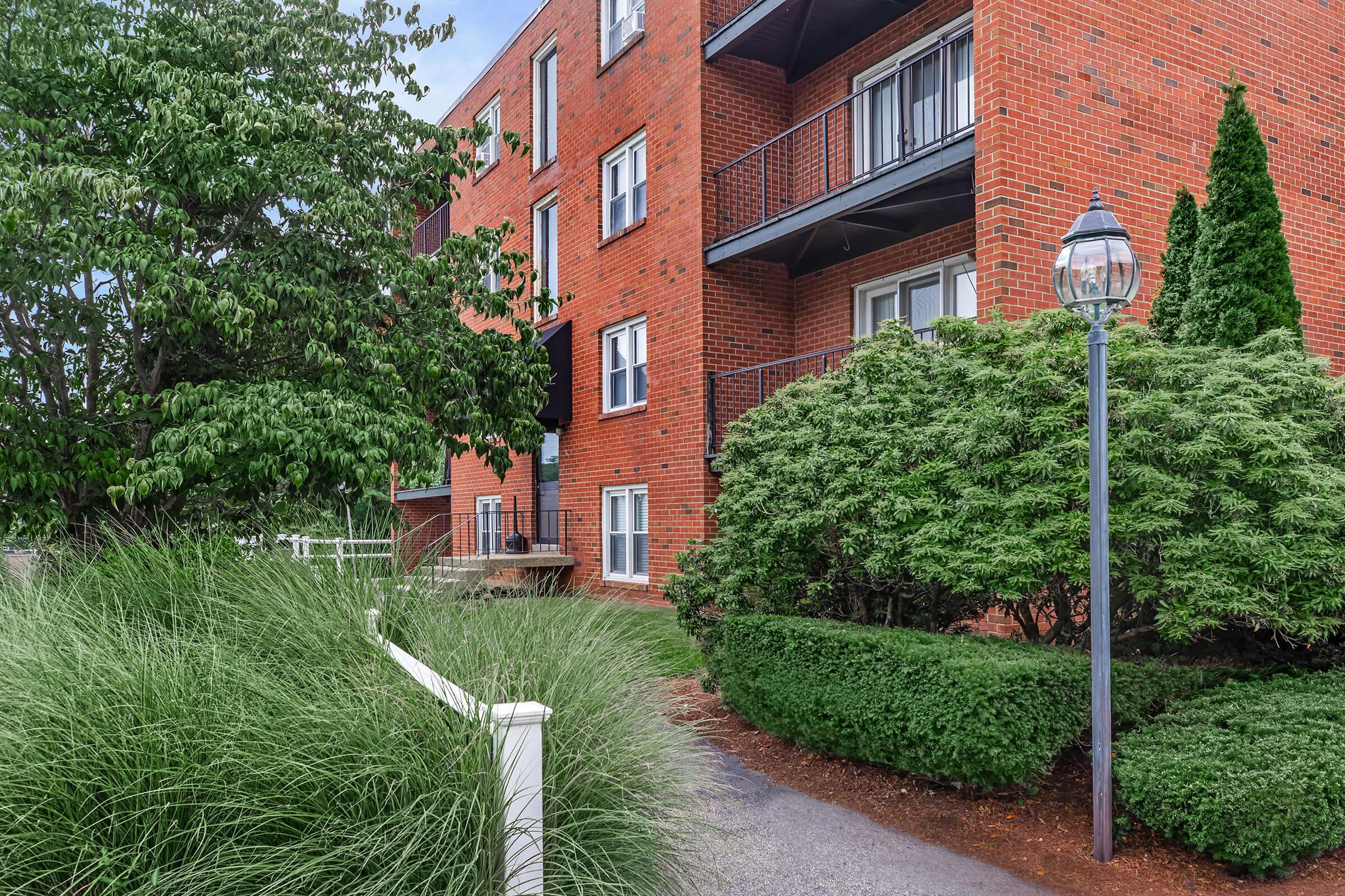 A brick apartment building with balconies, surrounded by lush greenery. A pathway leads to the entrance, with bushes and ornamental grass enhancing the outdoor space. A lamppost stands nearby, adding a classic touch to the scene. The sky is partially cloudy, suggesting a pleasant day.