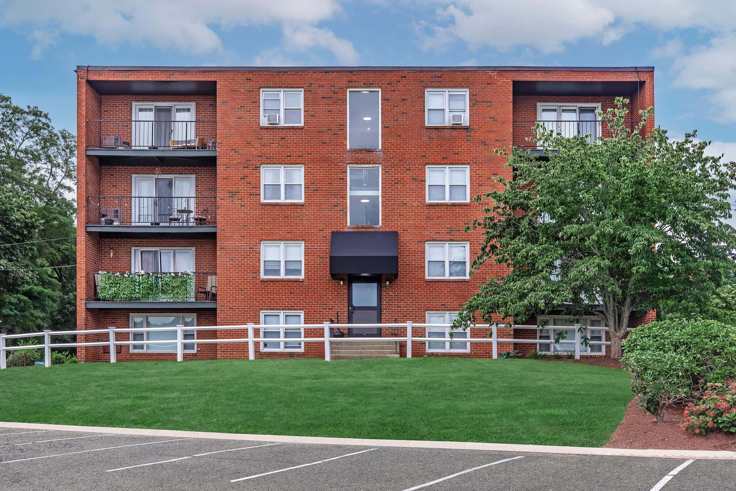 A red brick apartment building with four floors, featuring multiple balconies and large windows. The landscaping includes a grassy area in front of the building and a tree to the right. The parking lot is visible in the foreground, with marked parking spaces. The sky is partly cloudy.