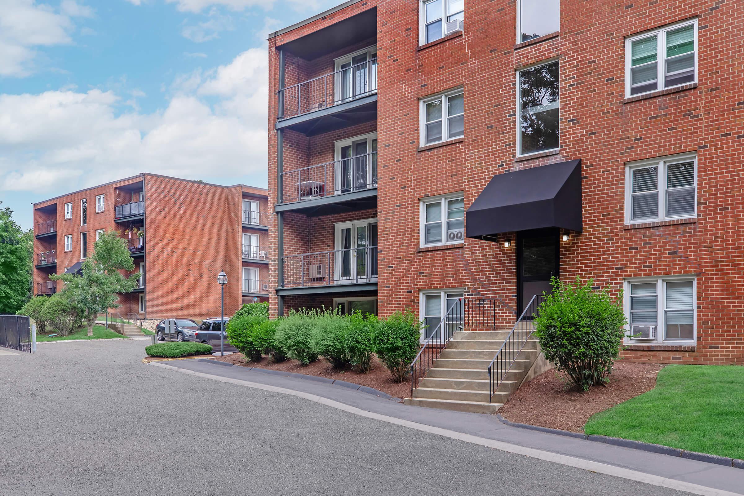 A multi-story brick apartment building with balconies, featuring a black awning over the entrance. In the foreground, there is a landscaped area with bushes, and a gravel road curving toward the building. Additional similar buildings are visible in the background under a partly cloudy sky.