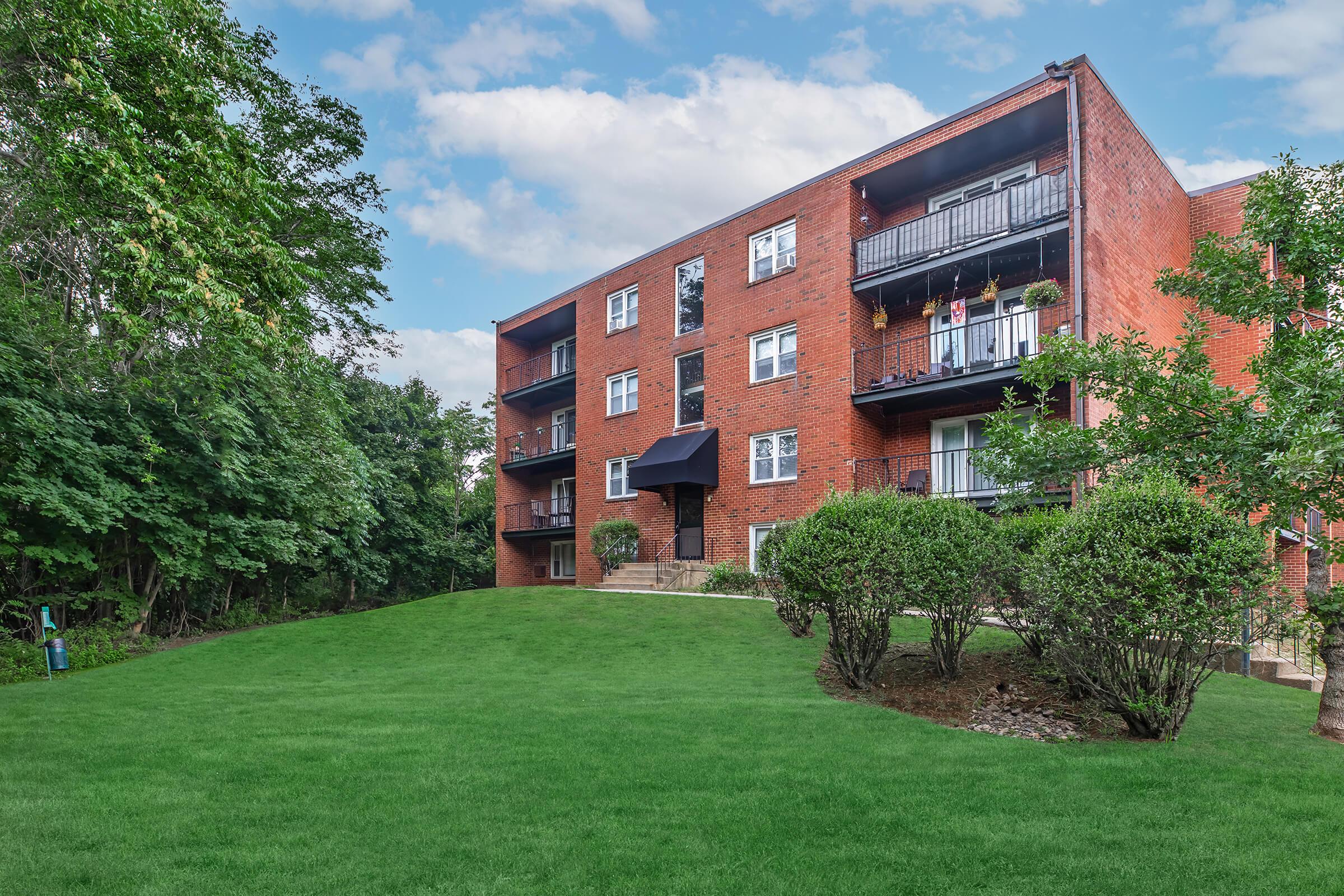 A brick apartment building with multiple balconies, surrounded by well-maintained greenery and a grassy lawn. The sky is partly cloudy, providing a pleasant backdrop to the residential setting. Trees are visible on one side, adding to the peaceful atmosphere.