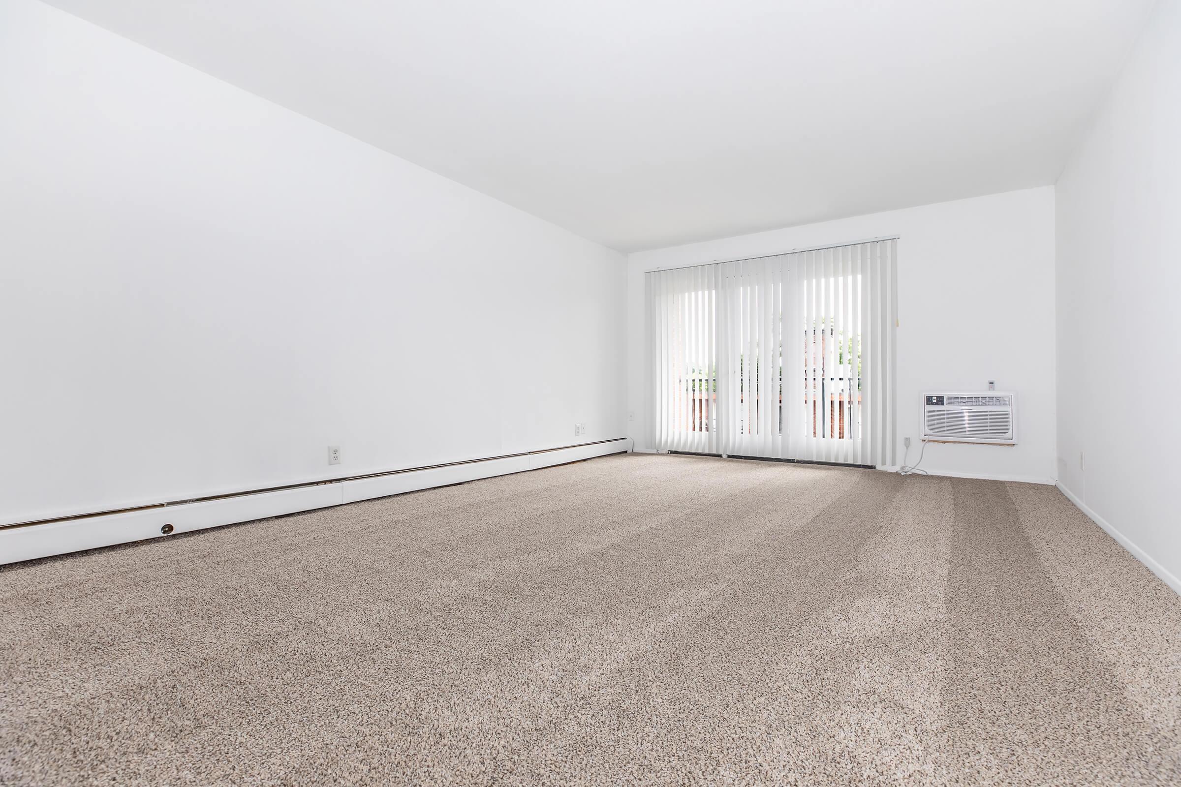 A spacious, empty room with light beige carpeting, painted white walls, and large sliding glass doors covered with vertical blinds, allowing natural light to enter. An air conditioning unit is mounted on the wall, enhancing the room's comfort.