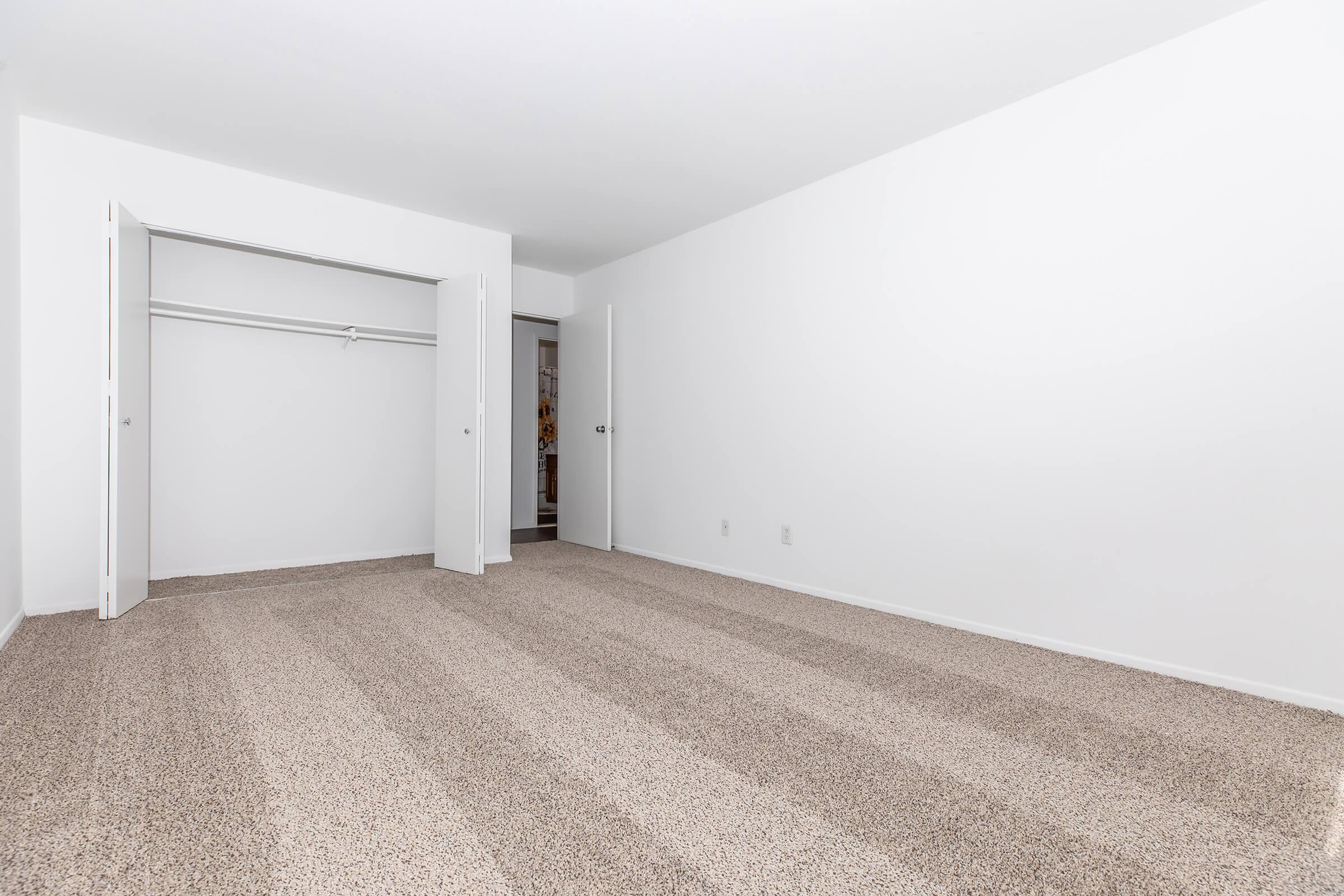 A spacious, empty room with beige carpet and white walls. There are two open doors leading to a closet, showing a simple and clean aesthetic. Natural light can be seen coming from an adjacent room through the doorway. The overall atmosphere is minimalistic and uncluttered.