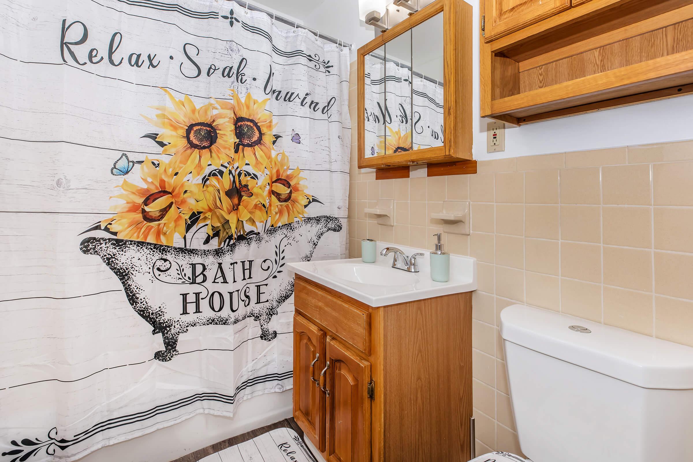 A bright bathroom featuring a sunflower-themed shower curtain with the text "Relax, Soak, Unwind." The space includes a wooden vanity with a sink, a wooden cabinet above, and a white toilet. The tile walls are a light beige color, creating a warm and inviting atmosphere.