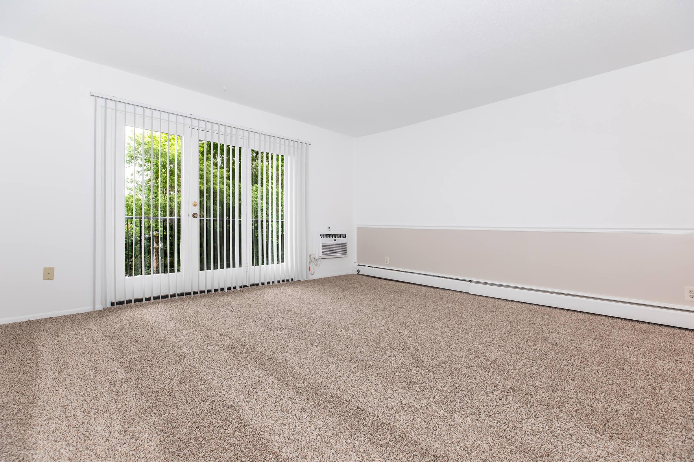 A vacant room with light beige carpet, featuring white walls and a decorative stripe near the bottom. The room has large sliding glass doors with vertical blinds that lead to an outdoor area, allowing natural light to enter. An air conditioning unit is visible along one wall.