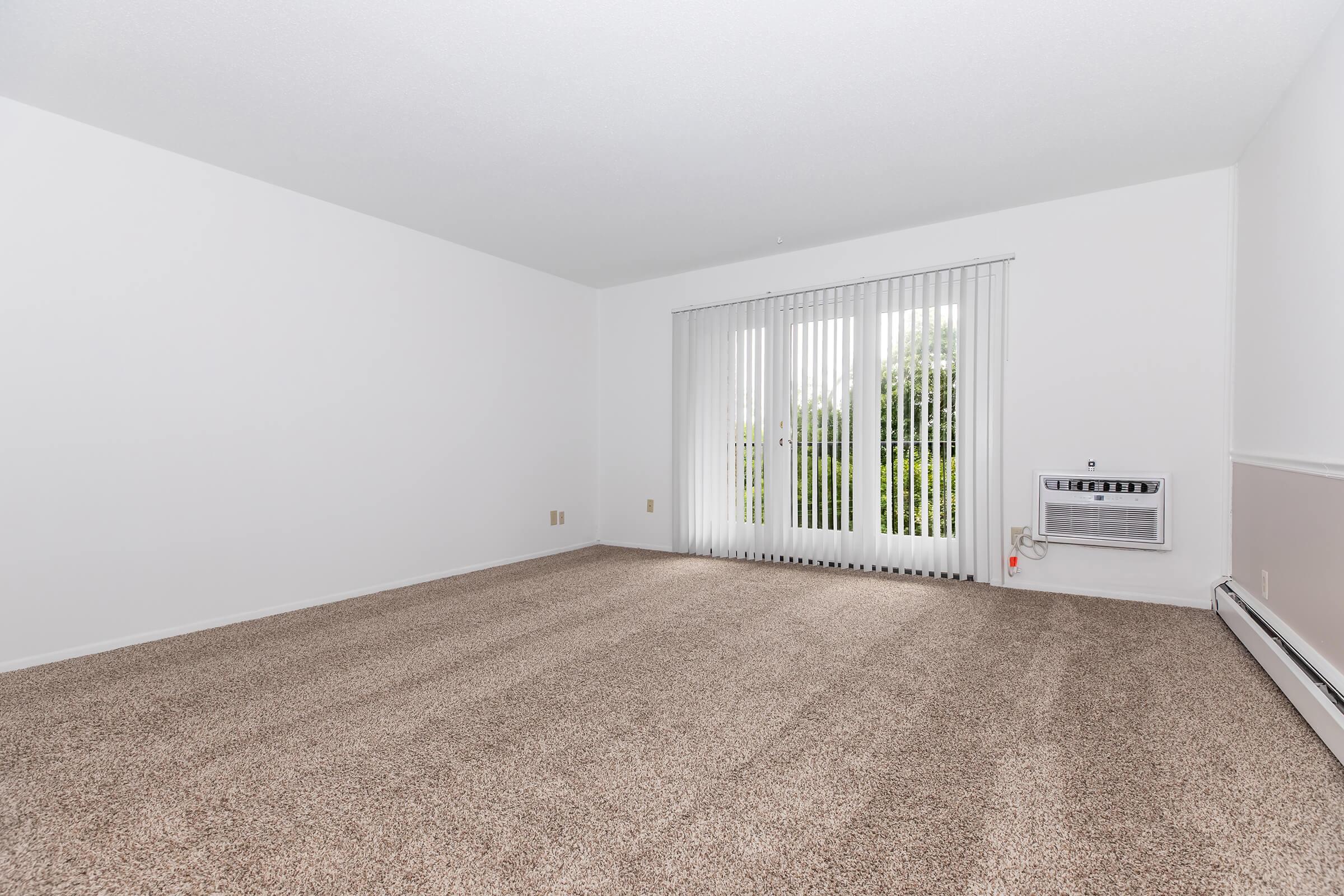Empty room with light beige carpet, white walls, and large windows covered with sheer vertical blinds. An air conditioning unit is visible on the right wall. The space is bright and airy, with natural light filtering in through the windows, suggesting a modern, minimalist interior design.