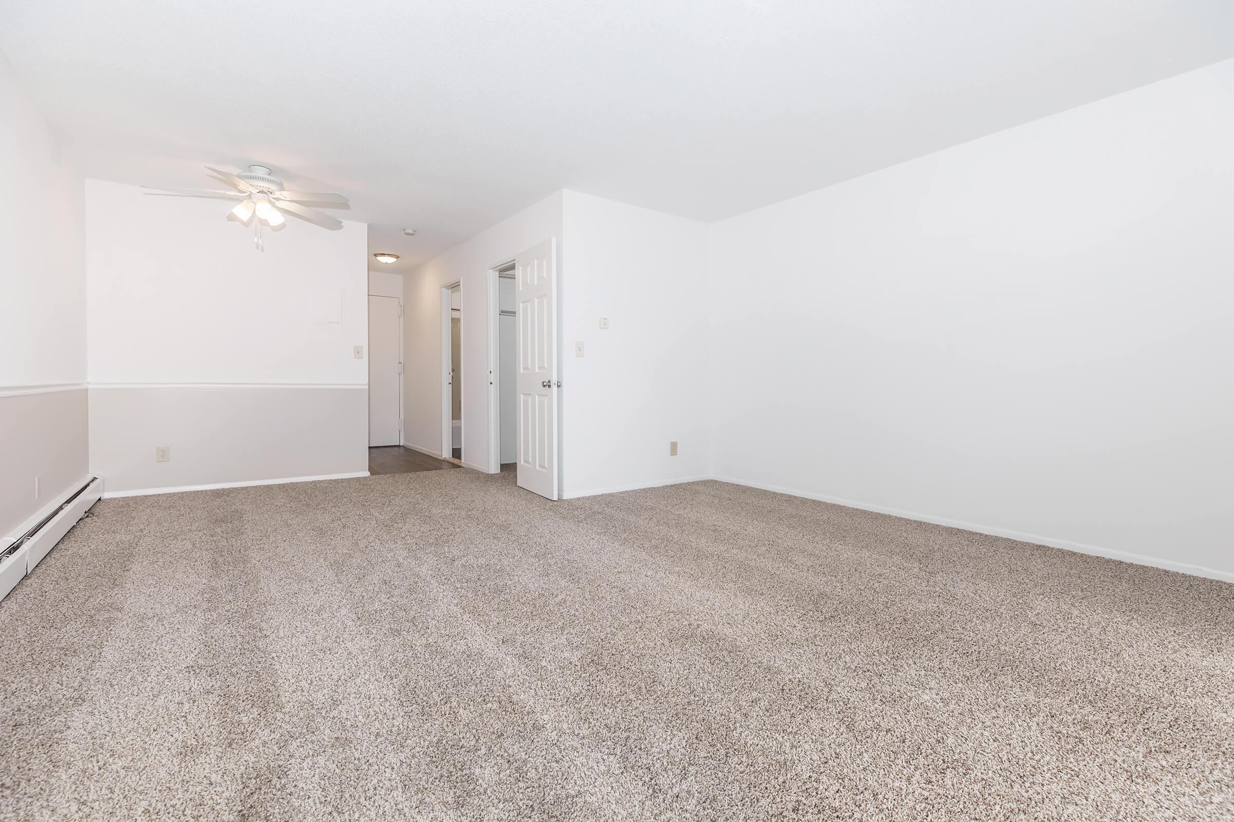 A spacious, empty living room featuring light-colored walls, plush carpet, and a ceiling fan. The room has a door leading to a hallway and an open layout, providing ample natural light and a clean, minimalistic appearance.