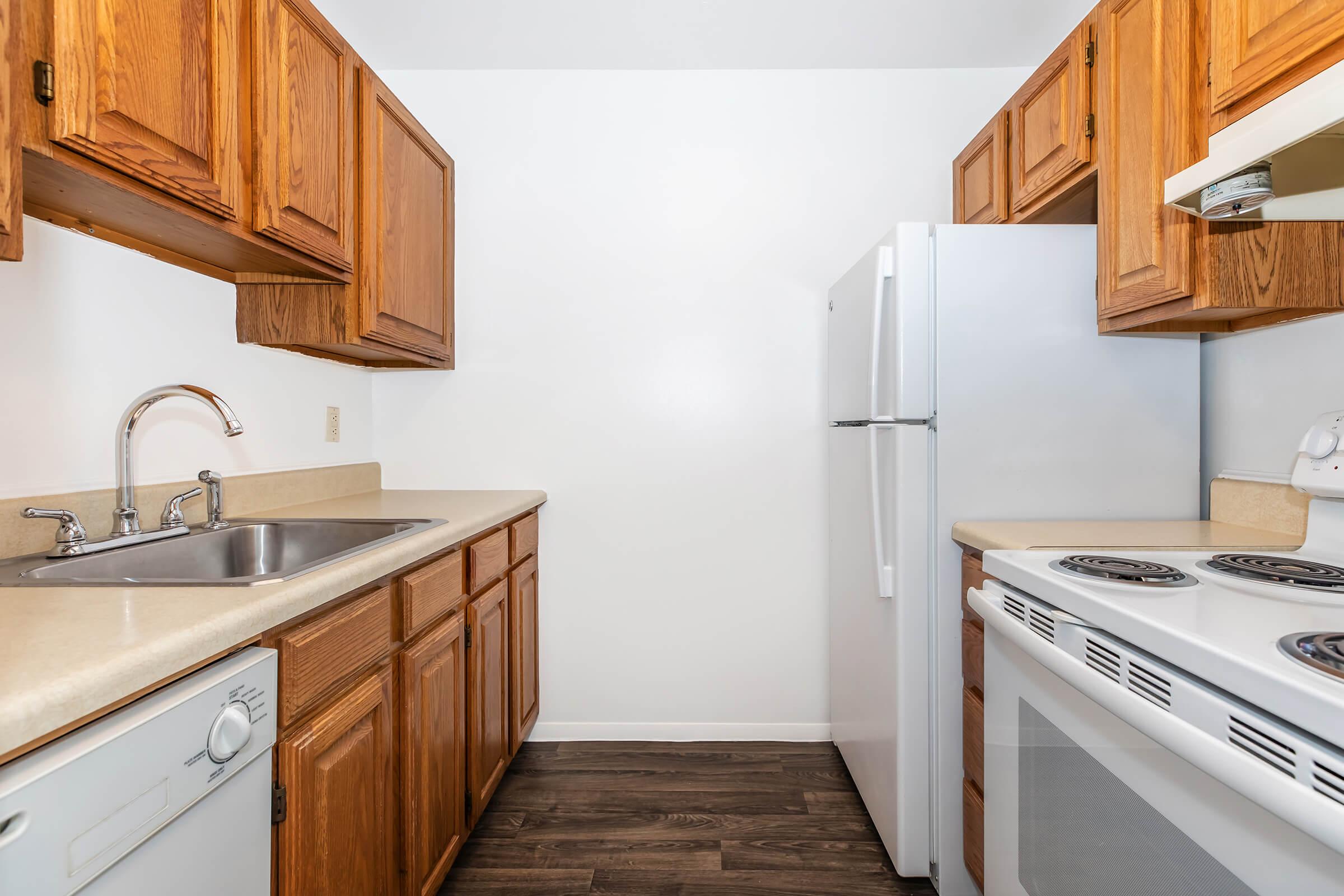 A bright kitchen featuring light wood cabinets, a double sink, a white refrigerator, and a stove with an oven. The countertop is beige, and the flooring is dark. The walls are plain white, creating a clean and inviting appearance.