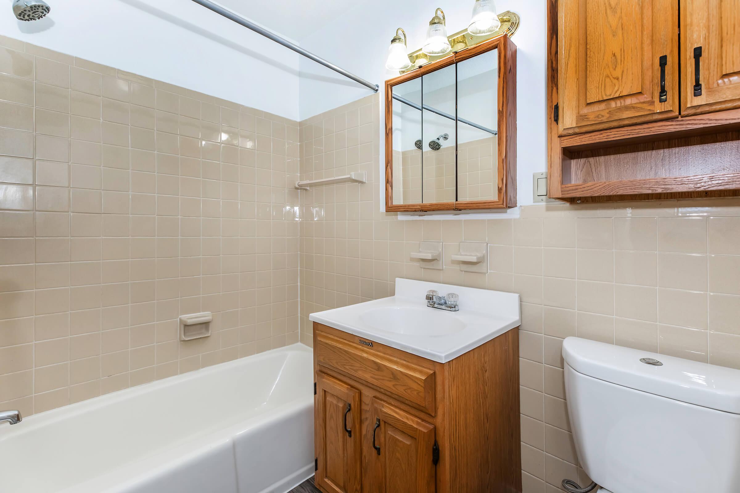 A clean bathroom featuring a bathtub with a showerhead, a wooden vanity with a white sink, a mirror above the sink, and a toilet. The walls are tiled in beige, and there's a wooden cabinet above the vanity for storage. The lighting includes four bulbs mounted above the mirror.