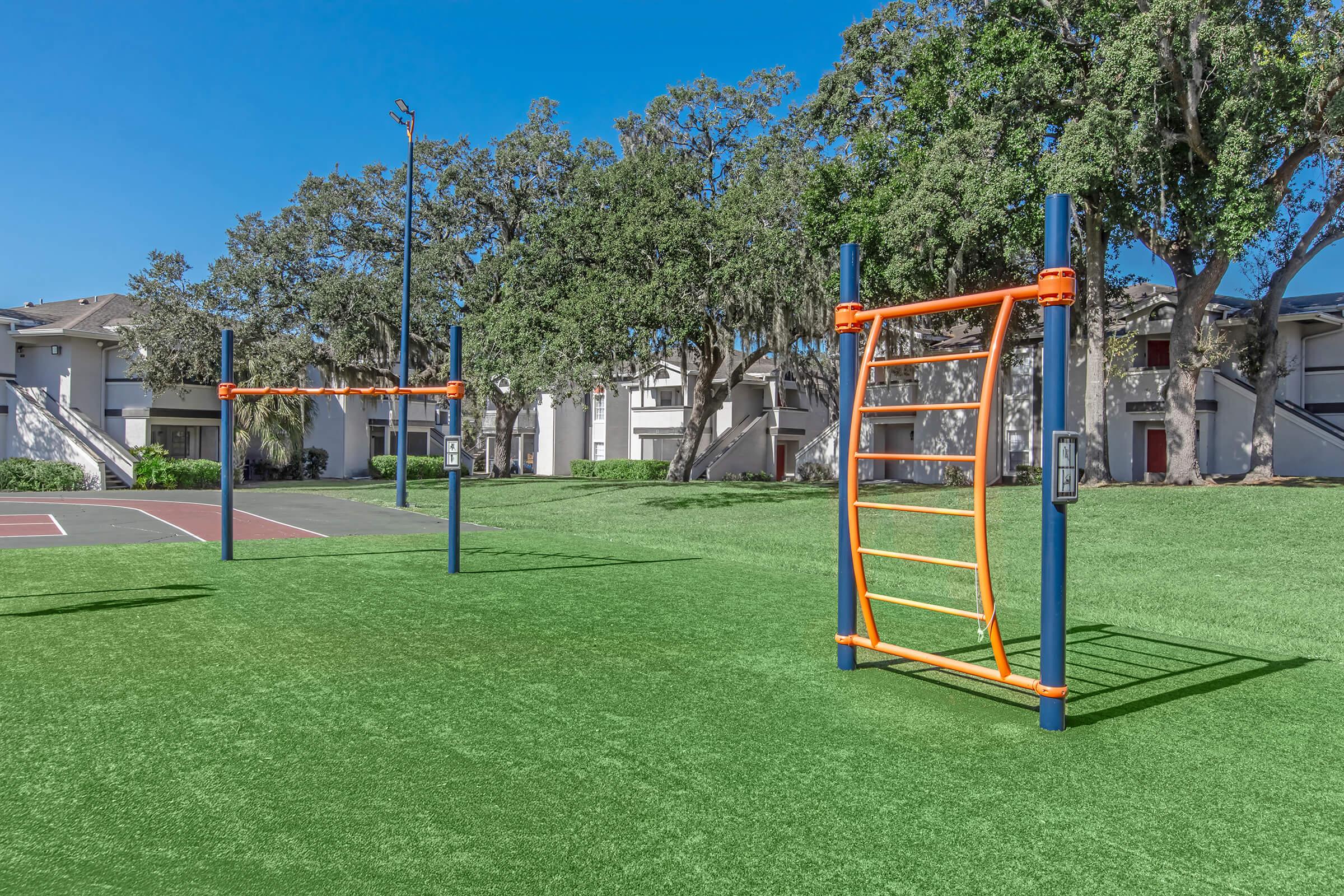 A bright outdoor fitness area featuring two sets of exercise equipment on green artificial turf, surrounded by trees and residential buildings in the background. The equipment includes bars for bodyweight exercises, with a clear blue sky overhead.