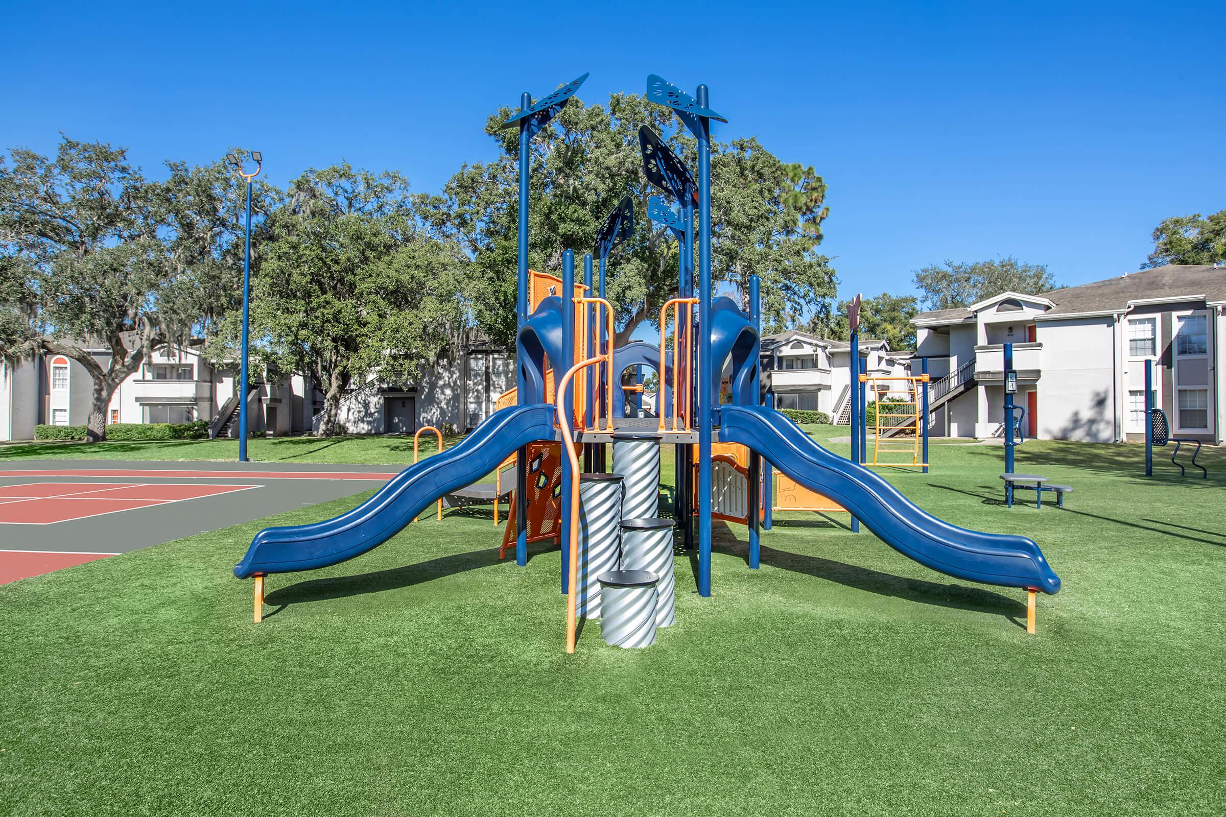 Colorful playground equipment with two blue slides, climbing structures, and a small tower surrounded by green grass. In the background, there are residential buildings and a basketball court with painted lines. The sky is clear and blue, creating a vibrant outdoor setting.