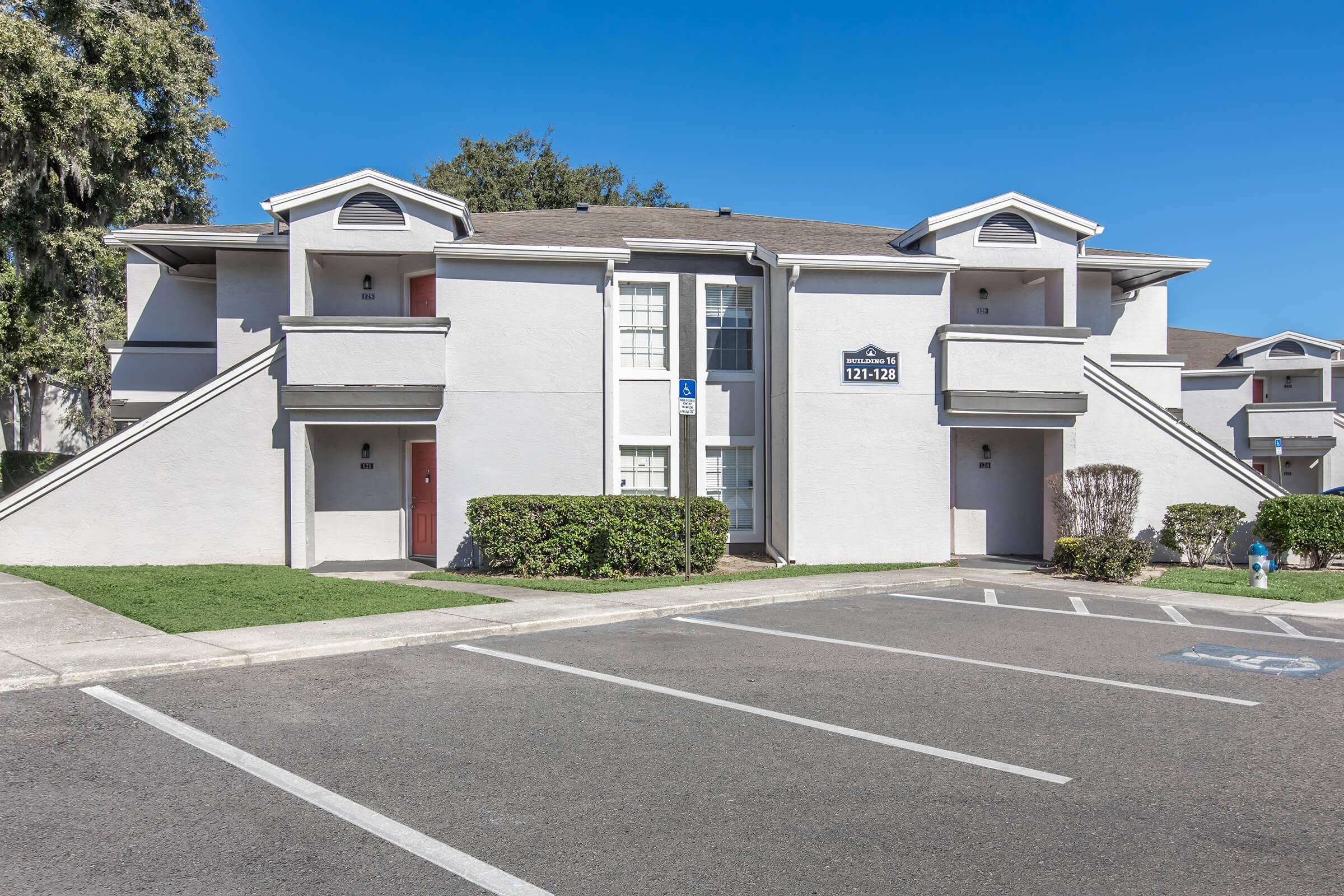 Two-story apartment building with a light gray exterior and a flat roof. The building features two sets of stairs leading to individual entrances, along with neatly trimmed bushes and a paved parking area in the foreground. Clear blue sky visible above.