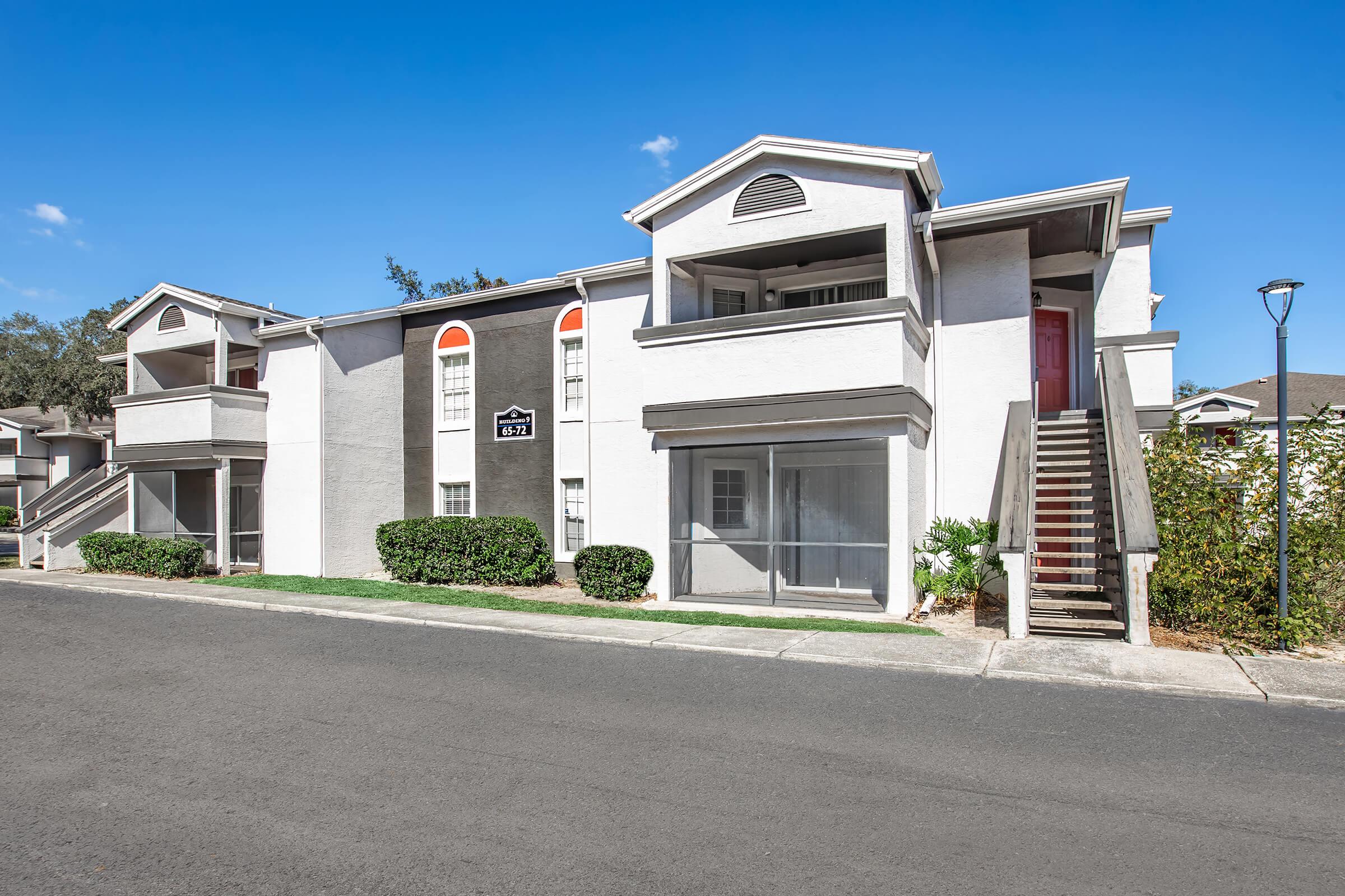 Exterior view of a two-story apartment building featuring a light gray façade with red accents. The building has several windows, a staircase leading to the second floor, and landscaped greenery along the front. The bright blue sky provides a contrasting backdrop.