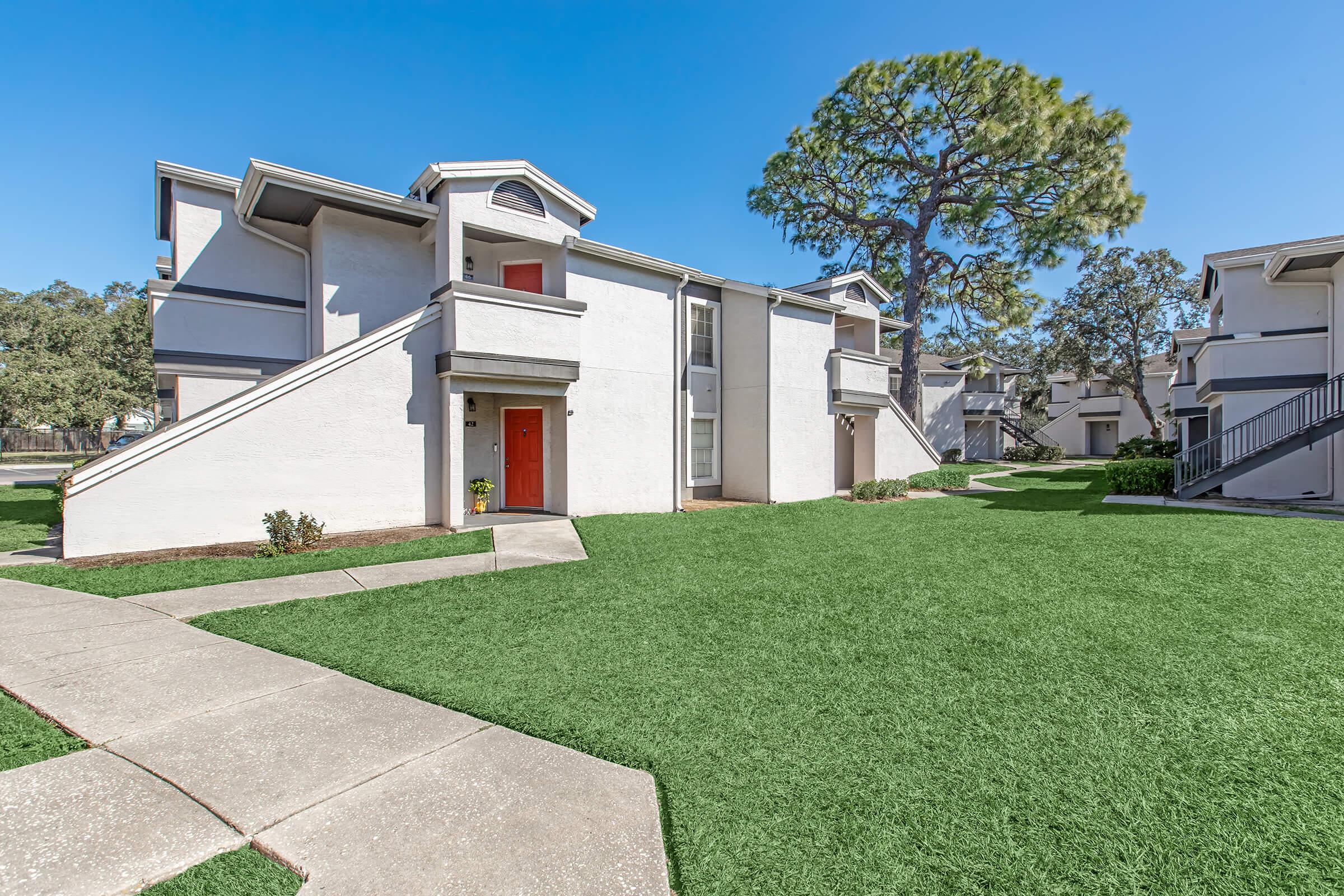 A two-story apartment building with a white exterior and red doors, surrounded by lush green grass and a large tree. The pathways are paved, leading to the entrances of the apartments, creating a welcoming outdoor space. Bright, clear blue skies complete the scene.