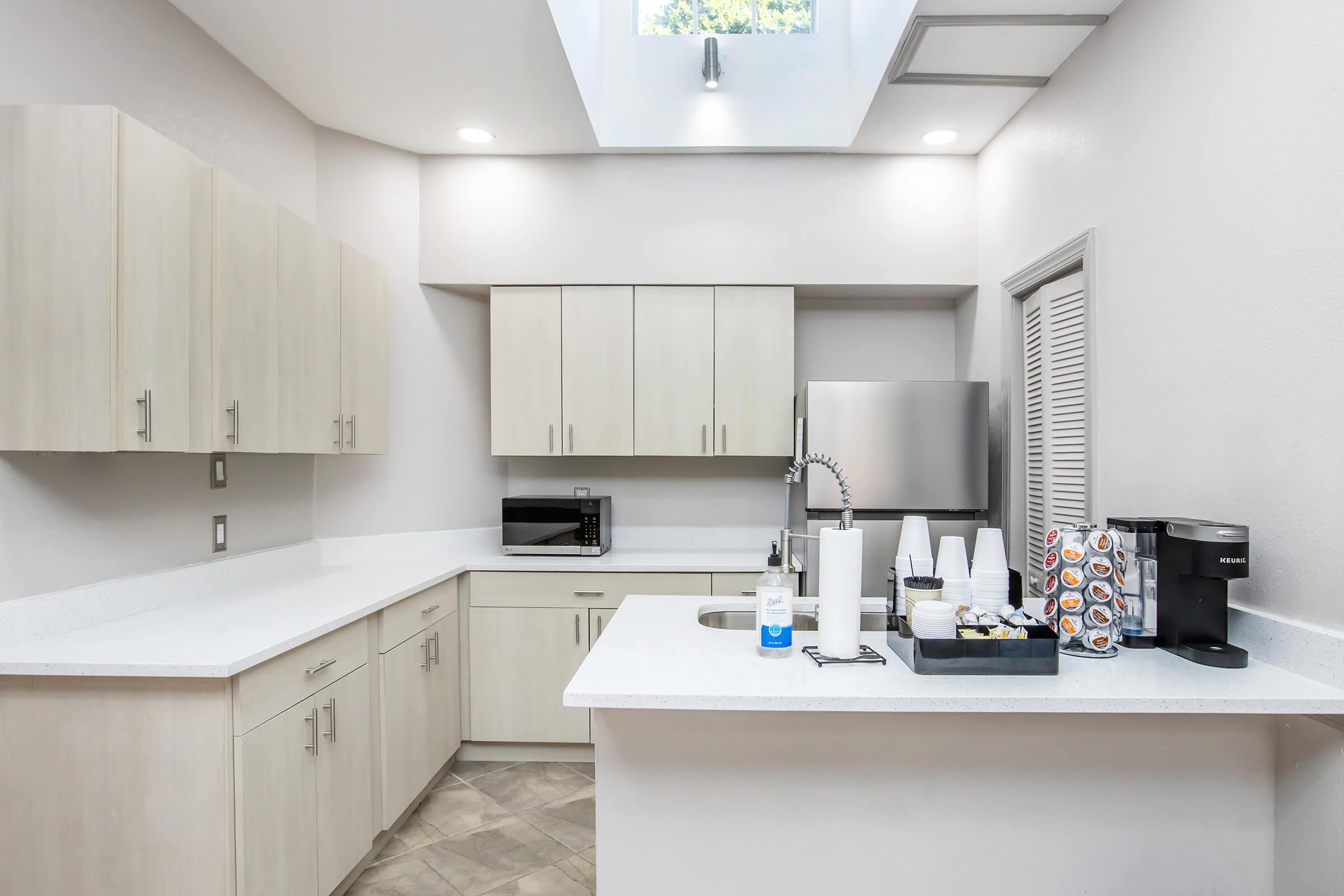 A modern kitchen featuring light-colored cabinets, a stainless steel refrigerator, a microwave, and a sink. The countertop is white with a coffee maker and a tray containing cups and other items. A skylight provides natural light, enhancing the clean and minimalist design of the space.