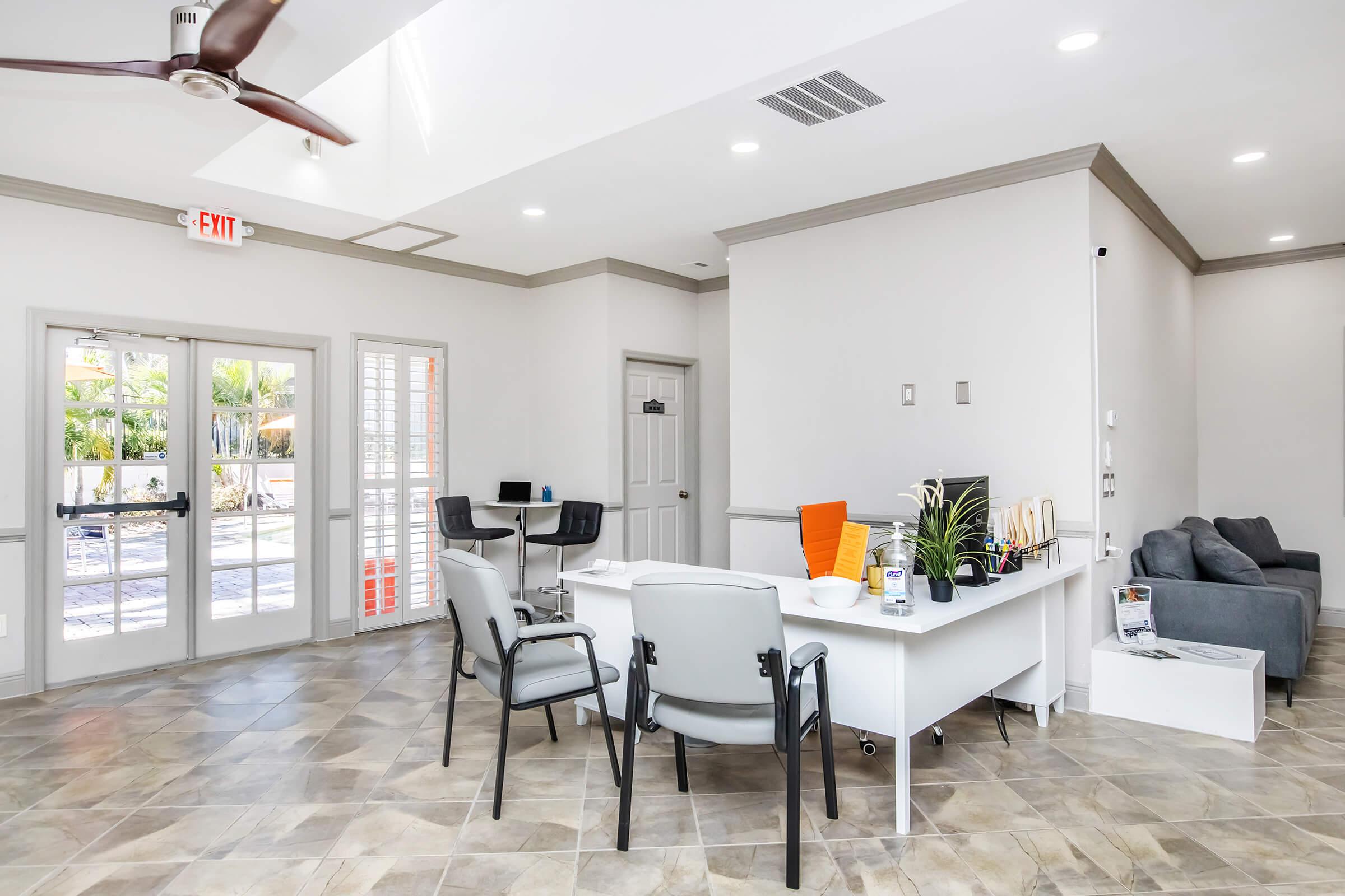 A modern office reception area with a large window letting in natural light. There are grey chairs, a white desk with a computer and office supplies, and a grey couch against the wall. The walls are light-colored, and the flooring is tiled. A ceiling fan is visible, creating a bright, inviting atmosphere.