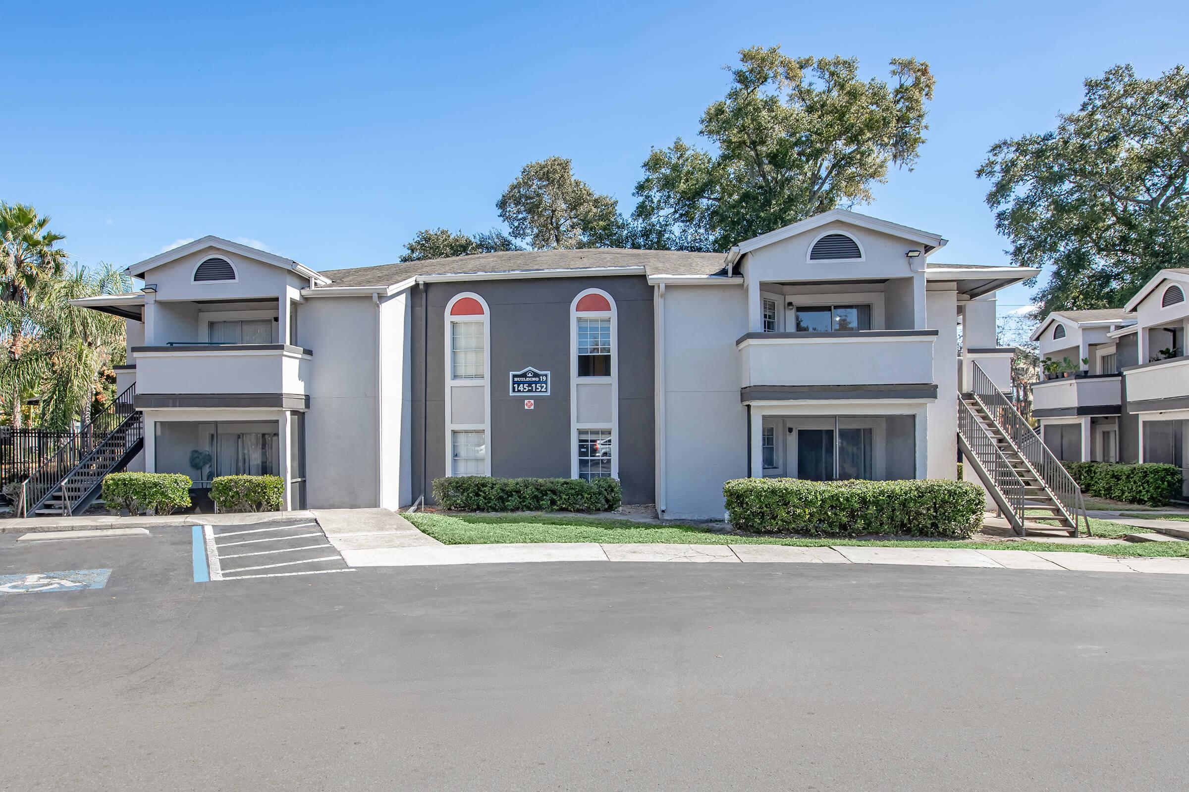 A two-story apartment building with a gray exterior. The building features multiple windows with red accents and white railings. There are neatly trimmed bushes in front and a paved pathway leading to the entrance. The area is sunny, surrounded by trees, and has a parking lot in view.