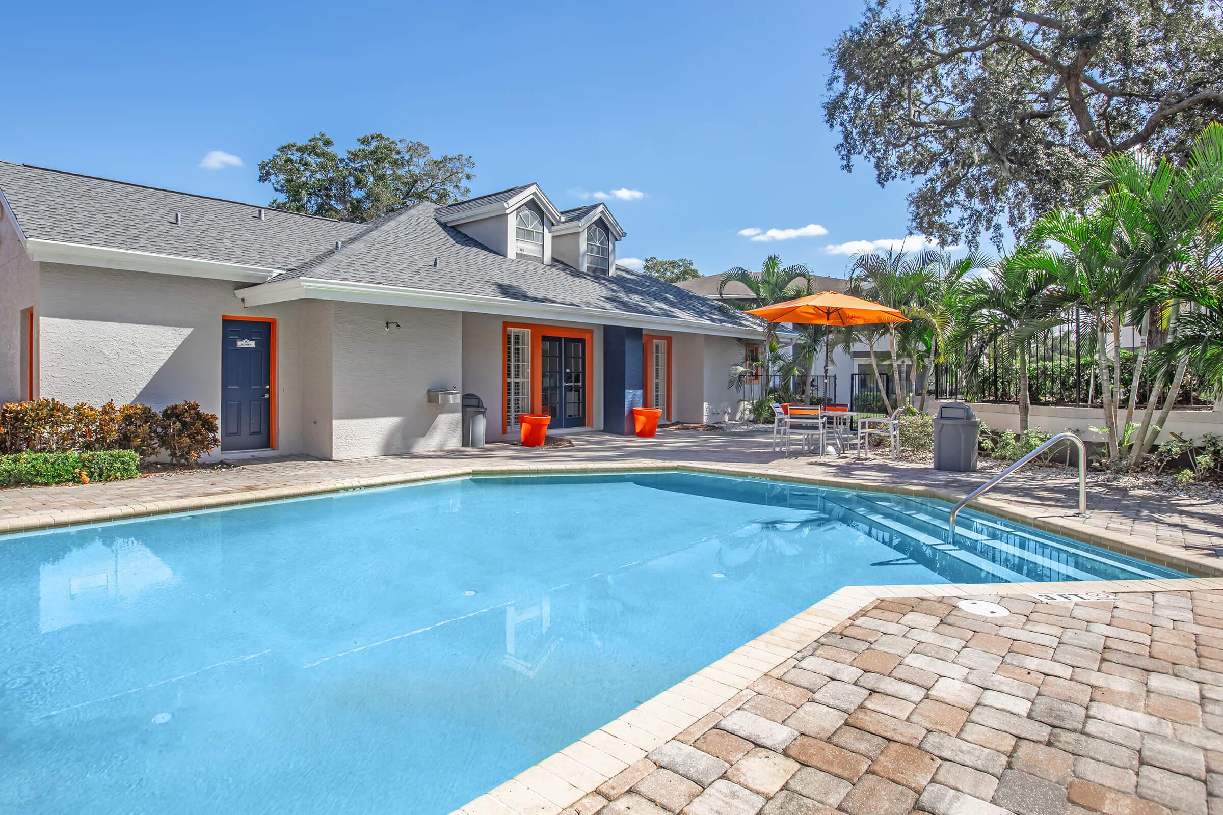 A sunny pool area featuring a blue swimming pool with a stone patio. Surrounding the pool are vibrant orange accents, including chairs and an umbrella. The backdrop includes a modern building with large windows and lush greenery, creating a relaxing outdoor space.