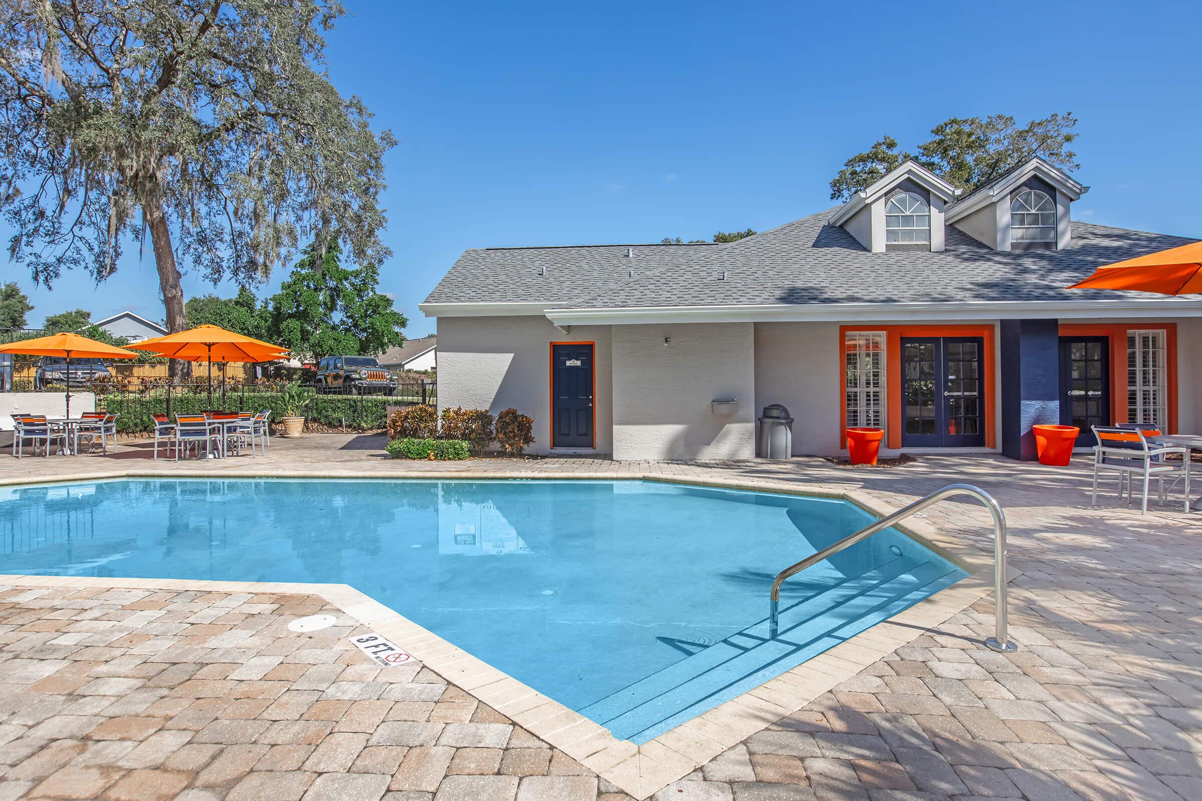 A bright outdoor pool area featuring a clear blue pool with a metal railing, surrounded by patio stones. In the background, a charming building with orange umbrellas and seating areas is visible, set against a clear blue sky and greenery.