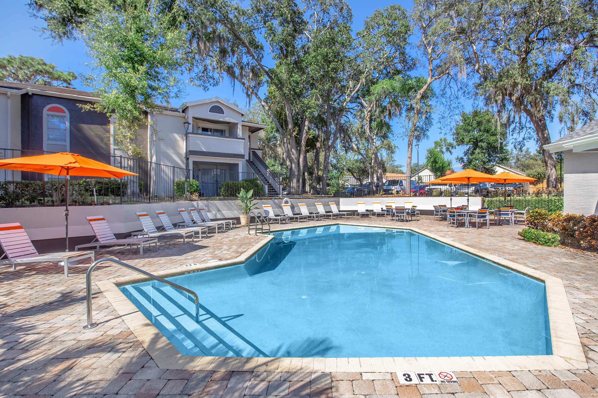 A vibrant outdoor pool area featuring a clear water pool with a shallow end marked at 3 feet. Surrounding the pool are orange umbrellas and lounge chairs, with a backdrop of lush trees and residential buildings. The area appears inviting and well-maintained, perfect for relaxation.