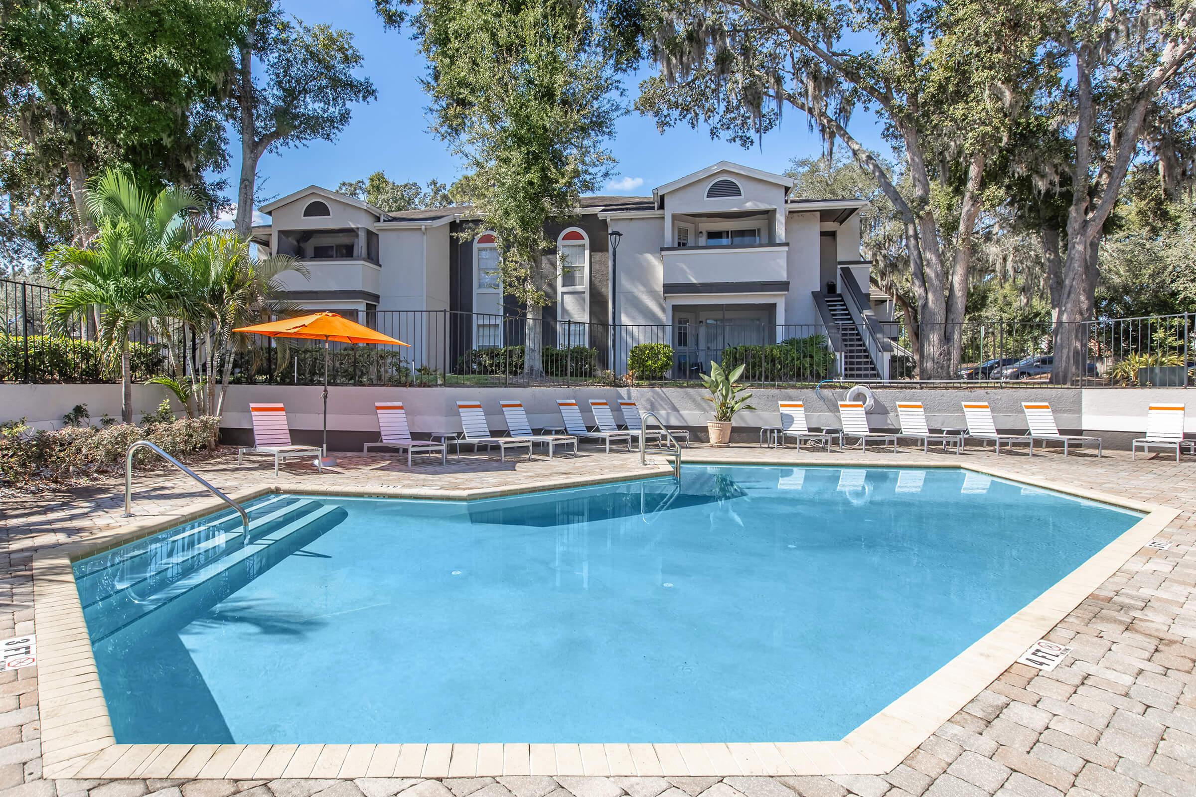 A well-maintained outdoor pool area featuring a clear blue water pool surrounded by lounge chairs. An orange umbrella provides shade, while lush trees and a multi-story apartment building are visible in the background, creating a relaxing atmosphere.