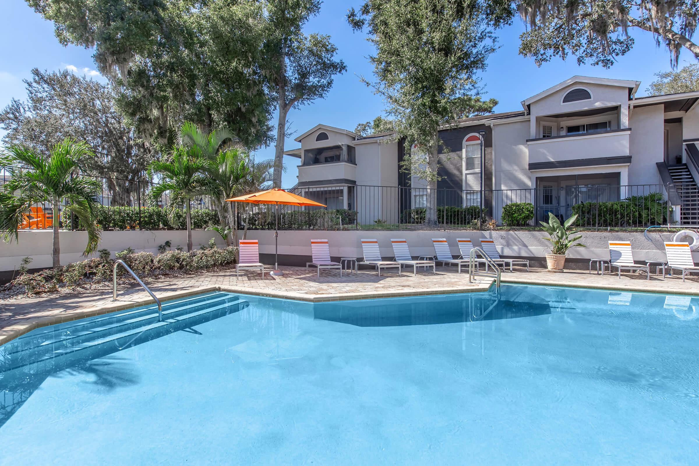 A serene swimming pool surrounded by lush greenery, with lounge chairs and an orange umbrella nearby. In the background, there are two-story buildings with balconies, set against a clear blue sky. The scene conveys a relaxing atmosphere ideal for leisure and enjoyment.