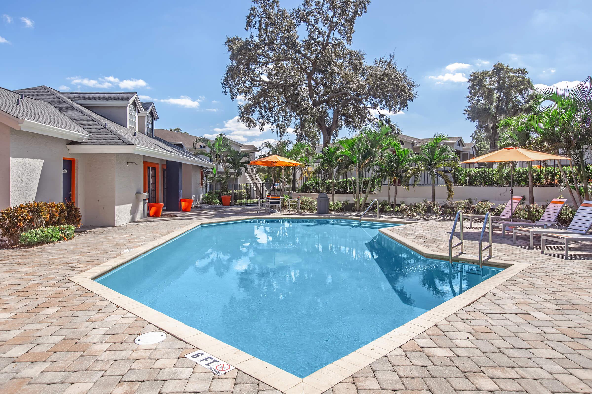 A clear swimming pool surrounded by patio tiles, with lounge chairs and orange umbrellas. Lush greenery and landscaping create a relaxing atmosphere, while nearby buildings showcase a residential setting. Bright sunlight enhances the tranquil vibe of the outdoor space.