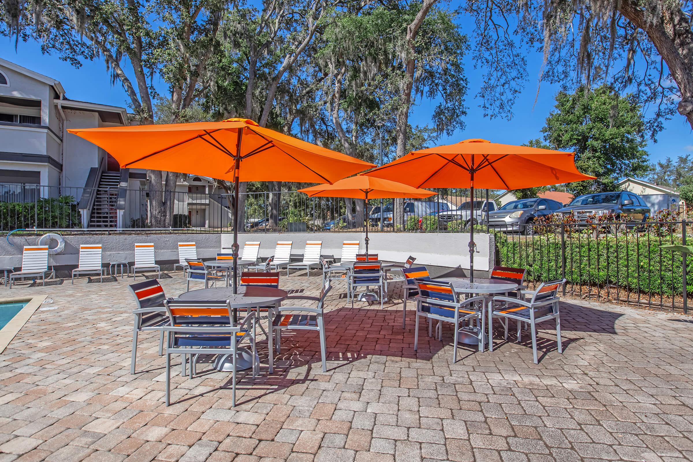Outdoor pool area featuring several round tables with orange umbrellas, surrounded by patterned cobblestone. In the background, there are apartment buildings and trees. The setting appears inviting and sunny, perfect for relaxation.