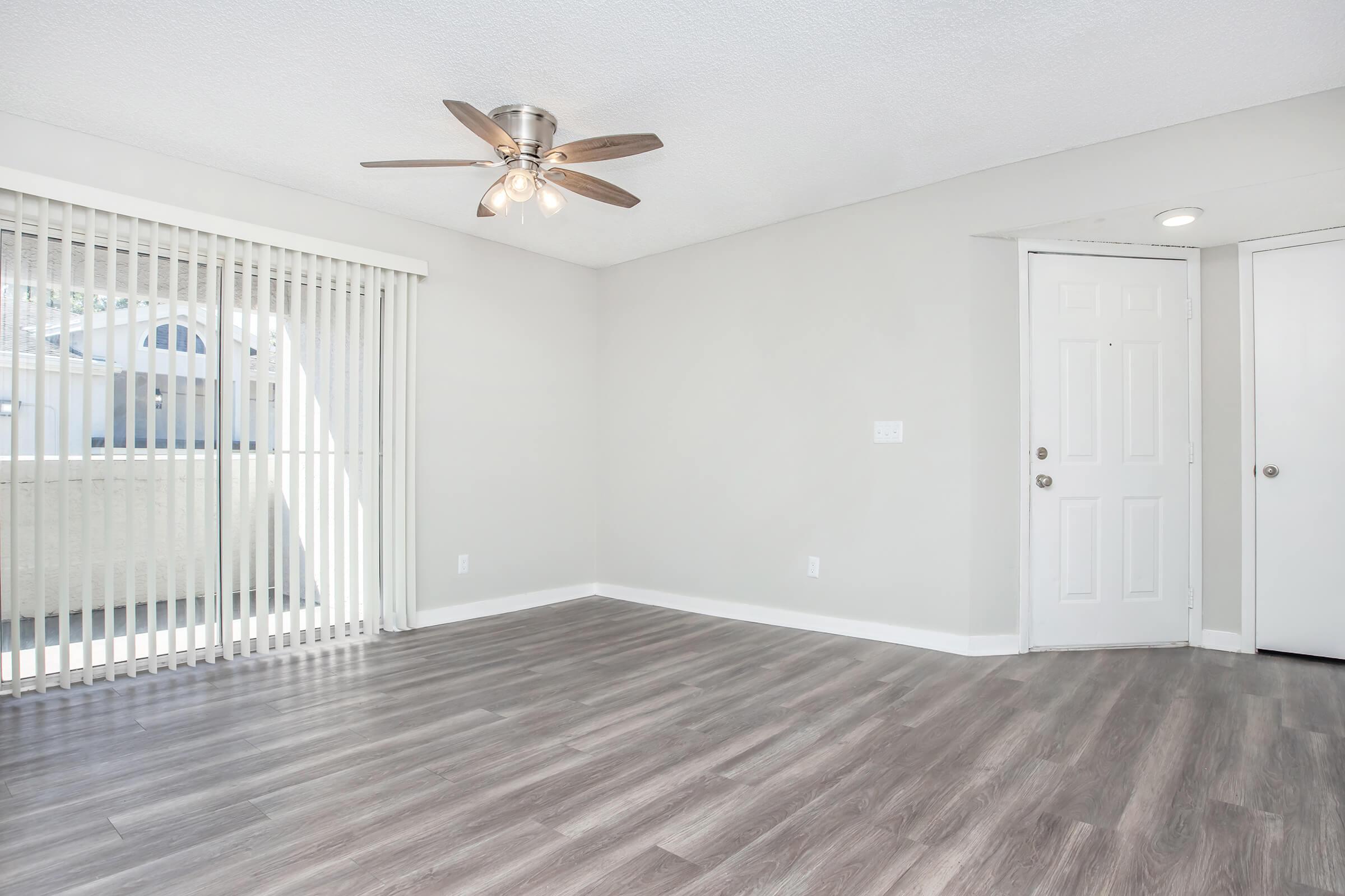 A bright, empty room featuring light gray walls and a modern ceiling fan. Large sliding glass doors with vertical blinds allow natural light. The floor is made of light wood laminate, and there is a white door on the right leading to another space.