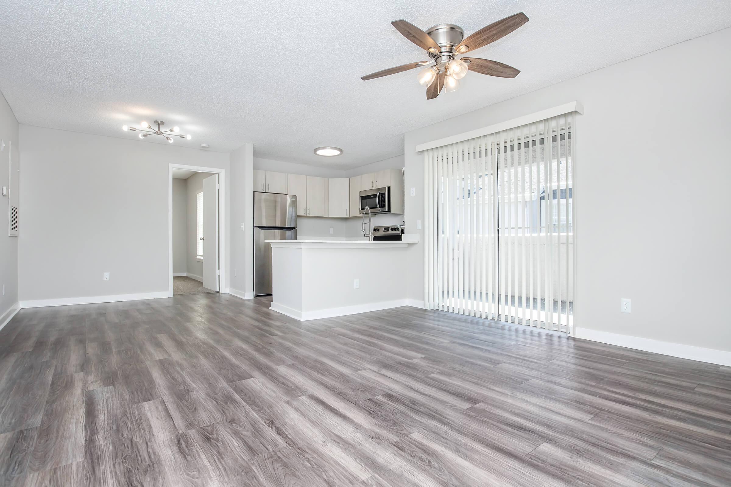 Spacious modern living area featuring wood-like flooring, a ceiling fan, and natural light from large sliding glass doors. The kitchen is partially visible with stainless steel appliances and an island, creating an open-concept space suitable for comfortable living and entertaining.