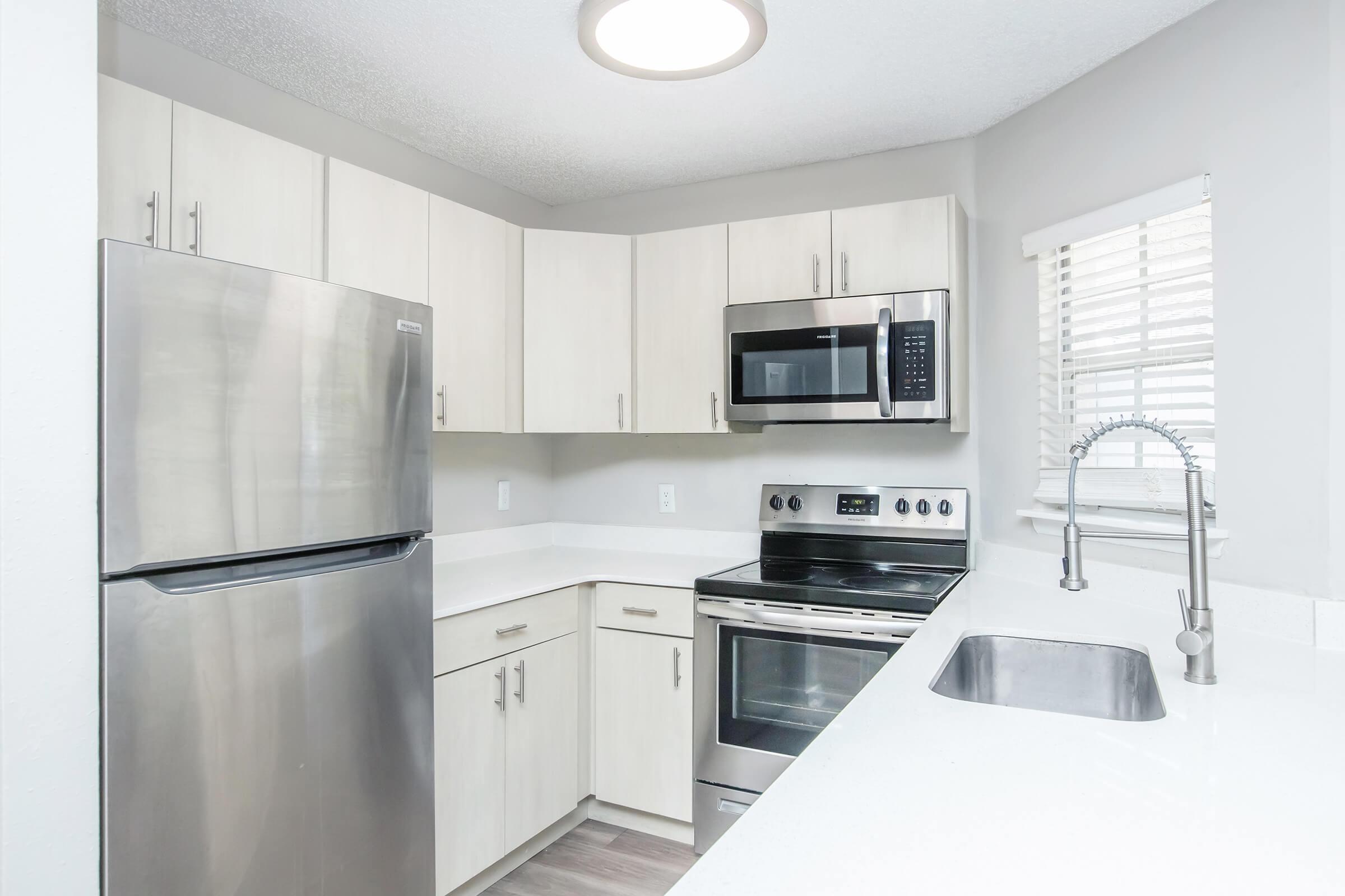 A modern kitchen featuring light-colored cabinets, stainless steel appliances including a refrigerator, microwave, and oven, and a white countertop. The sink is positioned beneath a window with blinds, and the space is well-lit with overhead lighting.