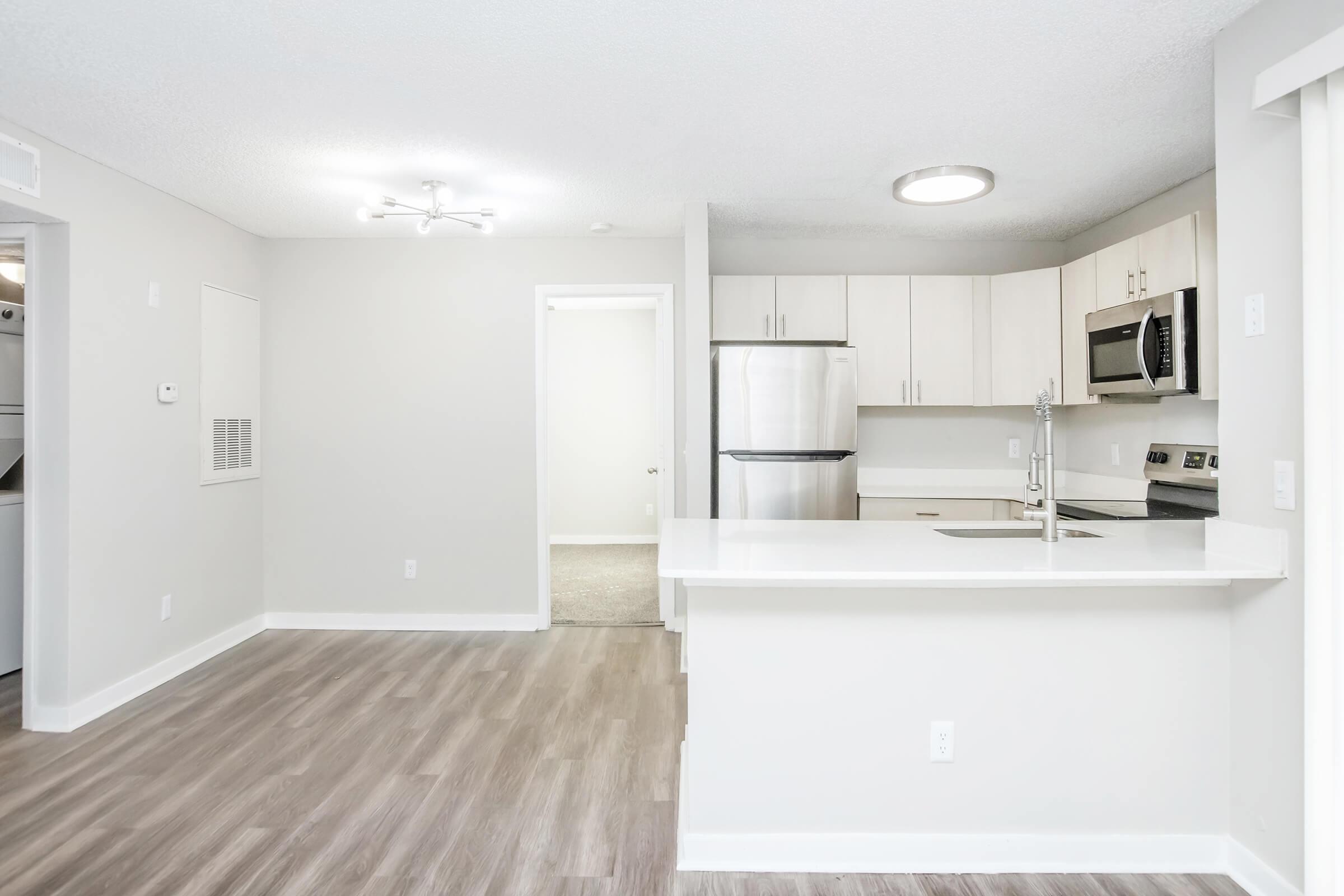 Modern apartment interior featuring an open kitchen with stainless steel appliances, a breakfast bar, and stylish cabinetry. The living area has warm wood-like flooring, bright LED lighting, and neutral walls, leading to a partially visible hallway and a door to another room.