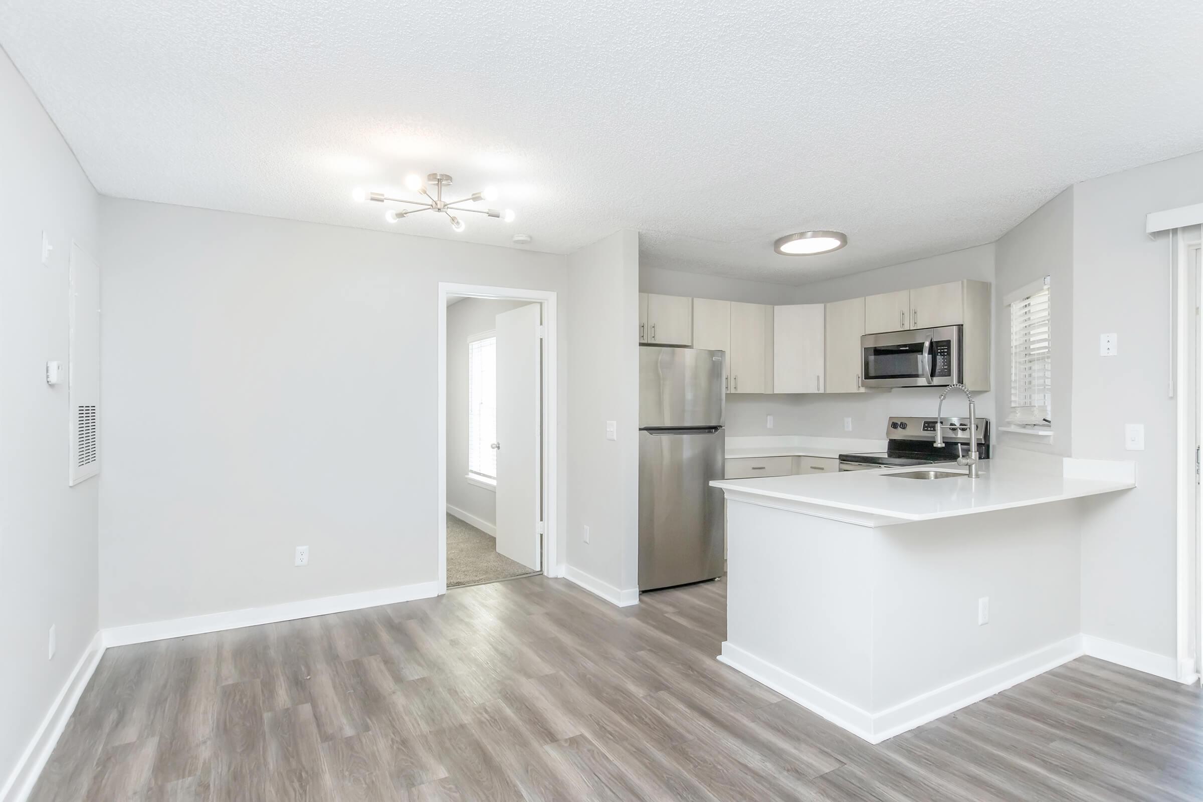 A modern kitchen with gray walls and wooden laminate flooring. It features stainless steel appliances including a refrigerator and microwave, a white countertop, and an open layout leading to a bright living area. Natural light enters through windows, enhancing the spacious feel of the room.