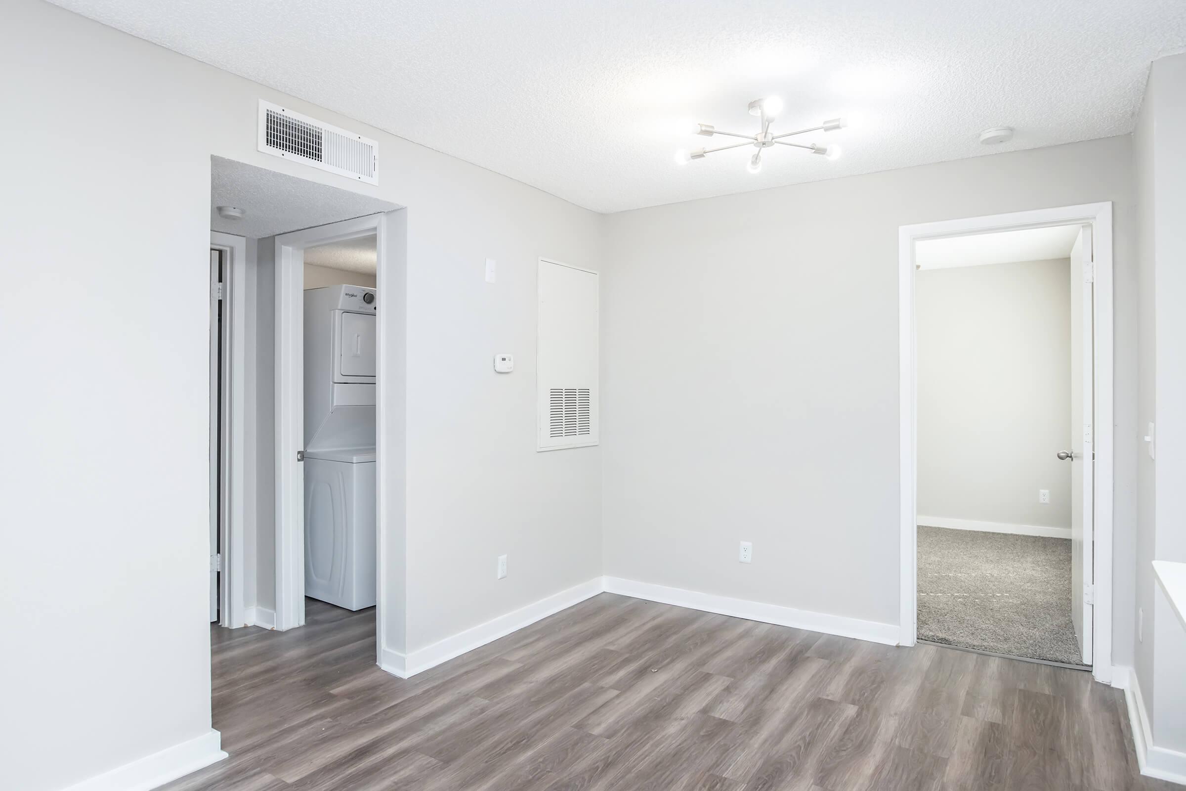 A bright, minimally decorated living space features light gray walls and wood-like flooring. To the left, a laundry area is visible through an open door, while a second door leads to another room. A modern ceiling light fixture illuminates the room, creating a spacious and inviting atmosphere.