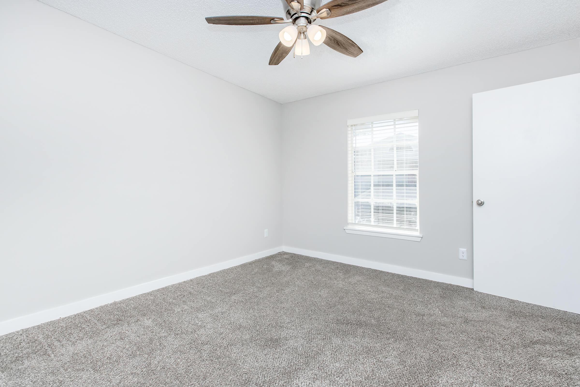 A well-lit empty room with light gray walls and carpet. It features a ceiling fan with three light bulbs, a window with white blinds, and a white door. The room has a clean, modern appearance and is ready for furniture arrangement or decoration.