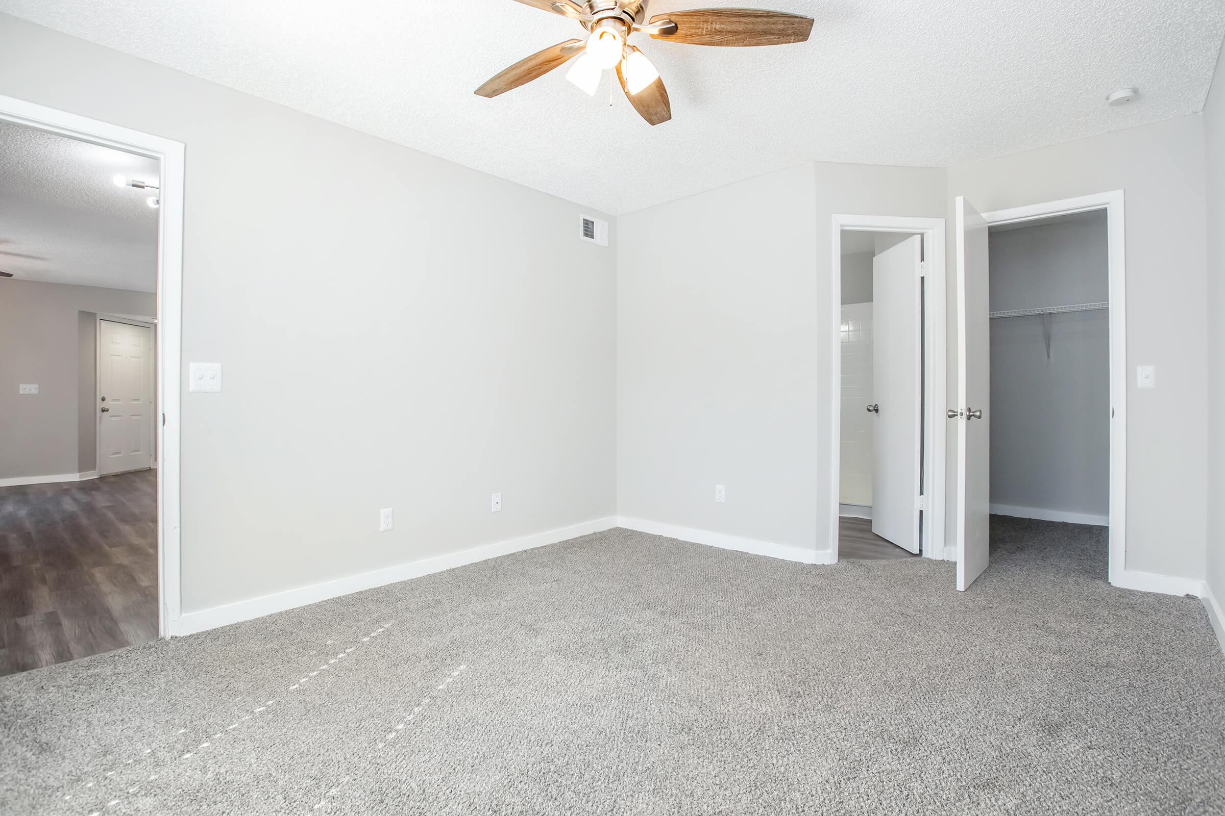 A clean, empty room with light gray walls and plush carpeting. A ceiling fan with wooden blades is overhead. Two doors lead to adjacent areas, one slightly ajar revealing a closet. Natural light brightens the space, creating an airy atmosphere.