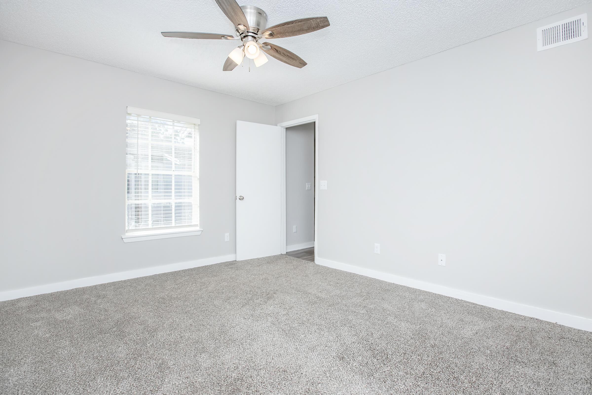 A well-lit, empty room featuring light gray walls and a ceiling fan with three bulbs. The floor is covered in soft beige carpet. One window, partially visible, allows natural light in, and a closed door leads to another room. The space is clean and ready for furniture.