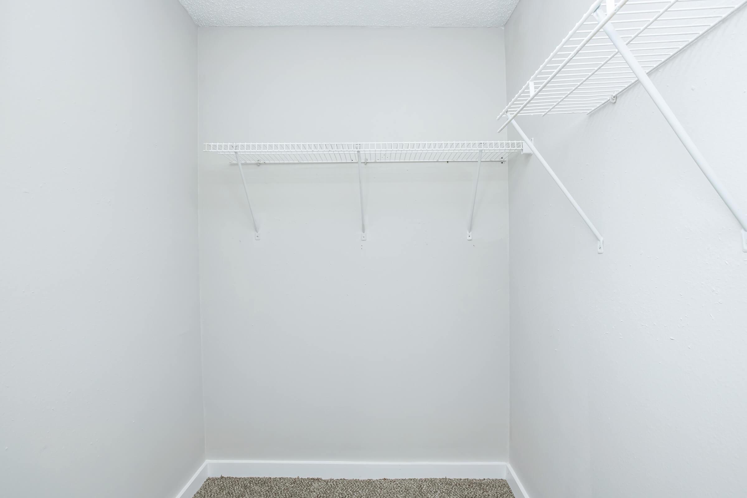 Empty closet with white walls and a beige carpet. Two metal wire shelves are mounted on the walls, with one on the left side and a partially visible one on the right. The space appears clean and organized, suitable for storage or organization.