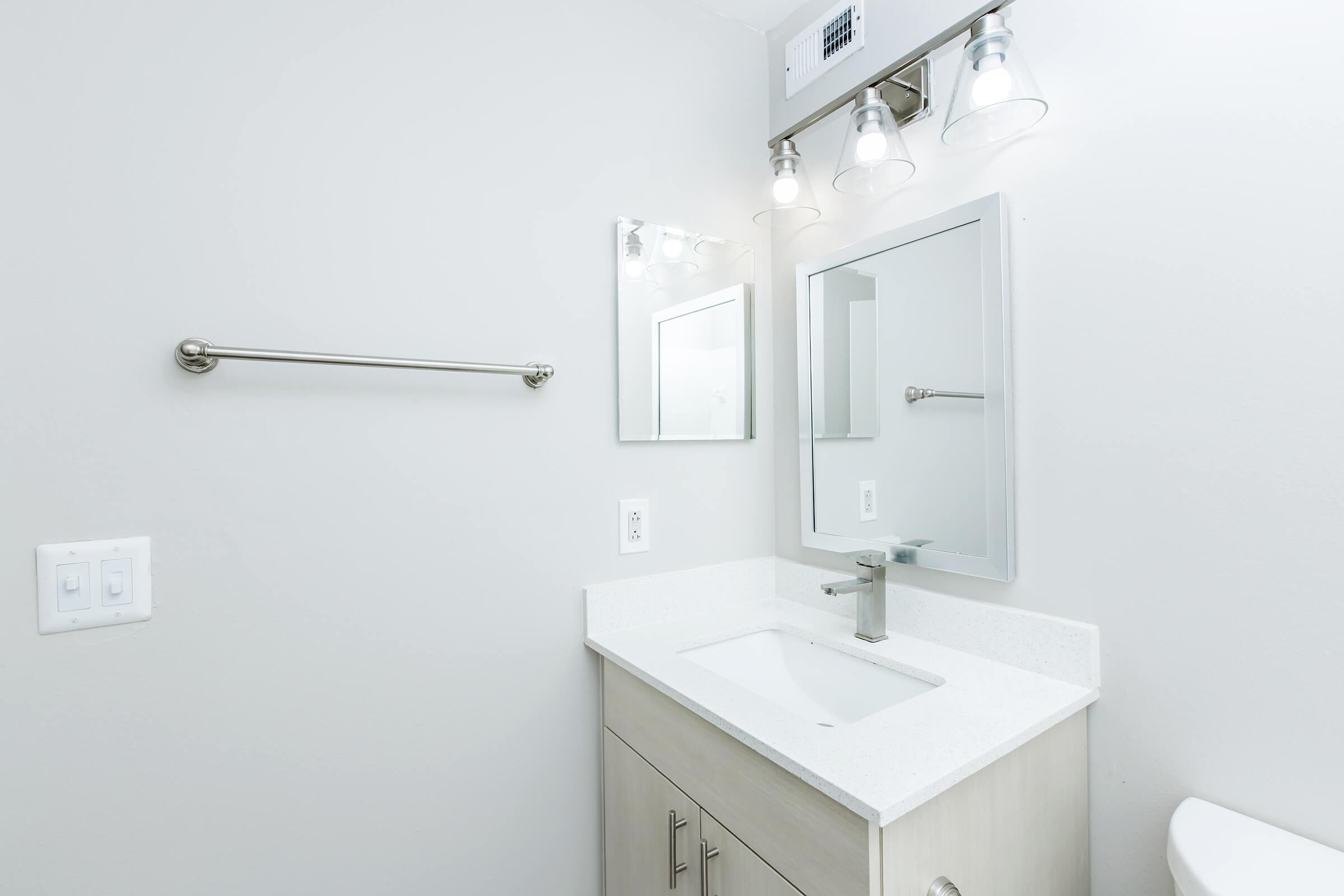 A modern bathroom featuring a white countertop sink, a wall-mounted mirror, three light fixtures above, a chrome towel bar, and a clean, minimalist design with light gray walls. The space is bright and well-lit, creating an inviting atmosphere.
